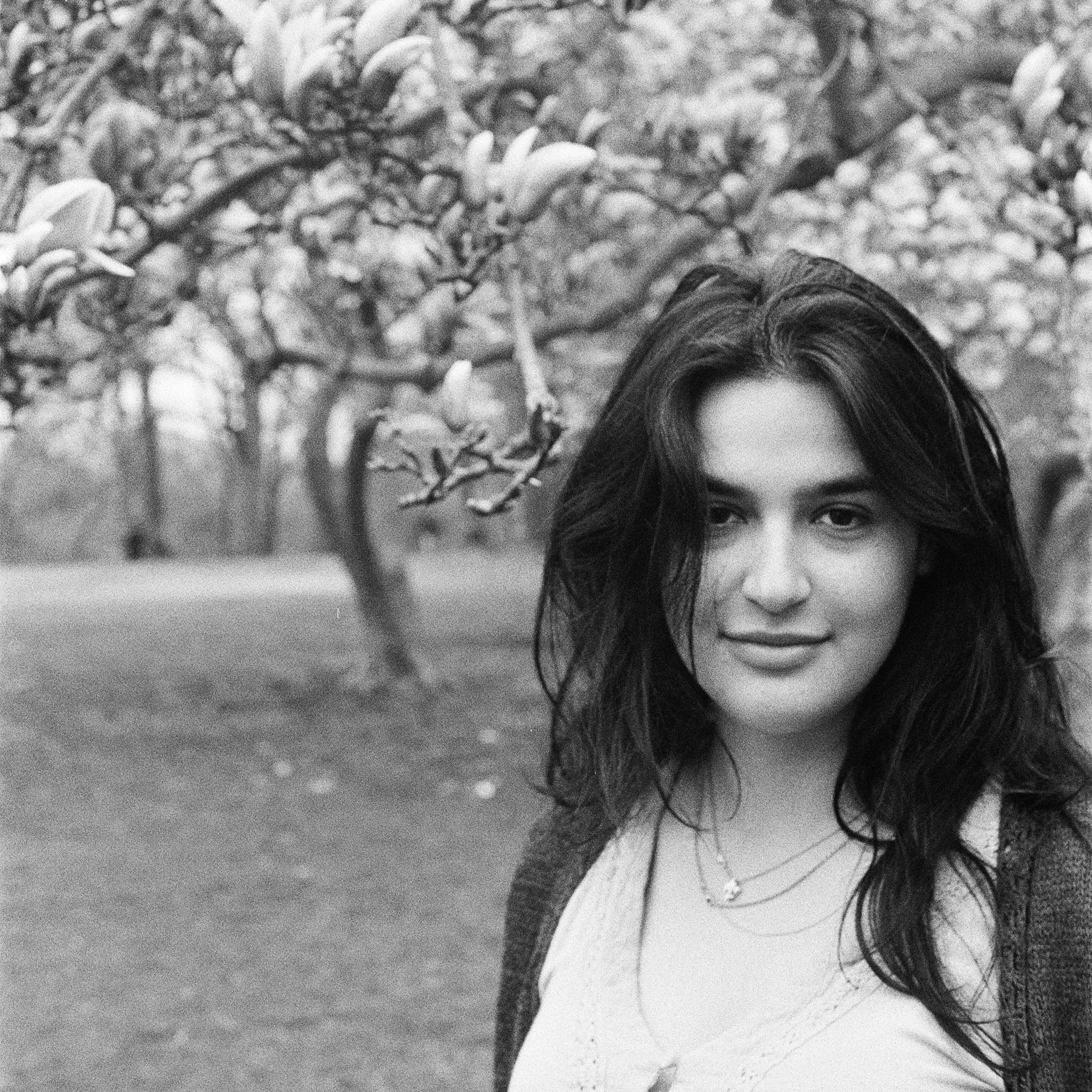 A black and white photo of a young woman with dark wavy hair standing outdoors near blossoming trees.