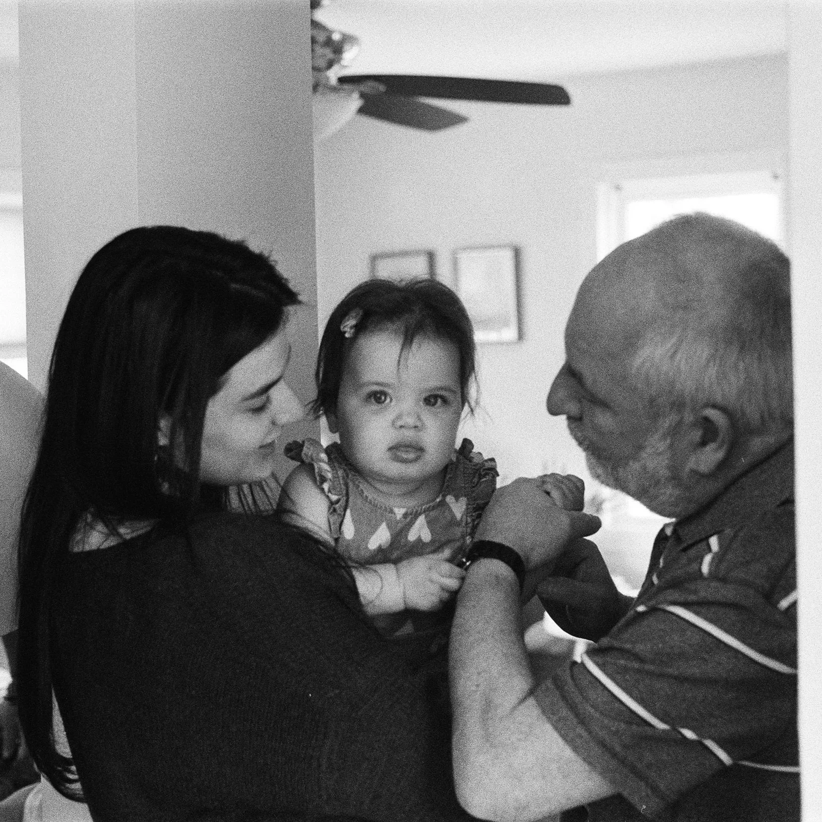 A woman holding a young girl with a serious expression, while an older man reaches out to touch the girl in a home setting.