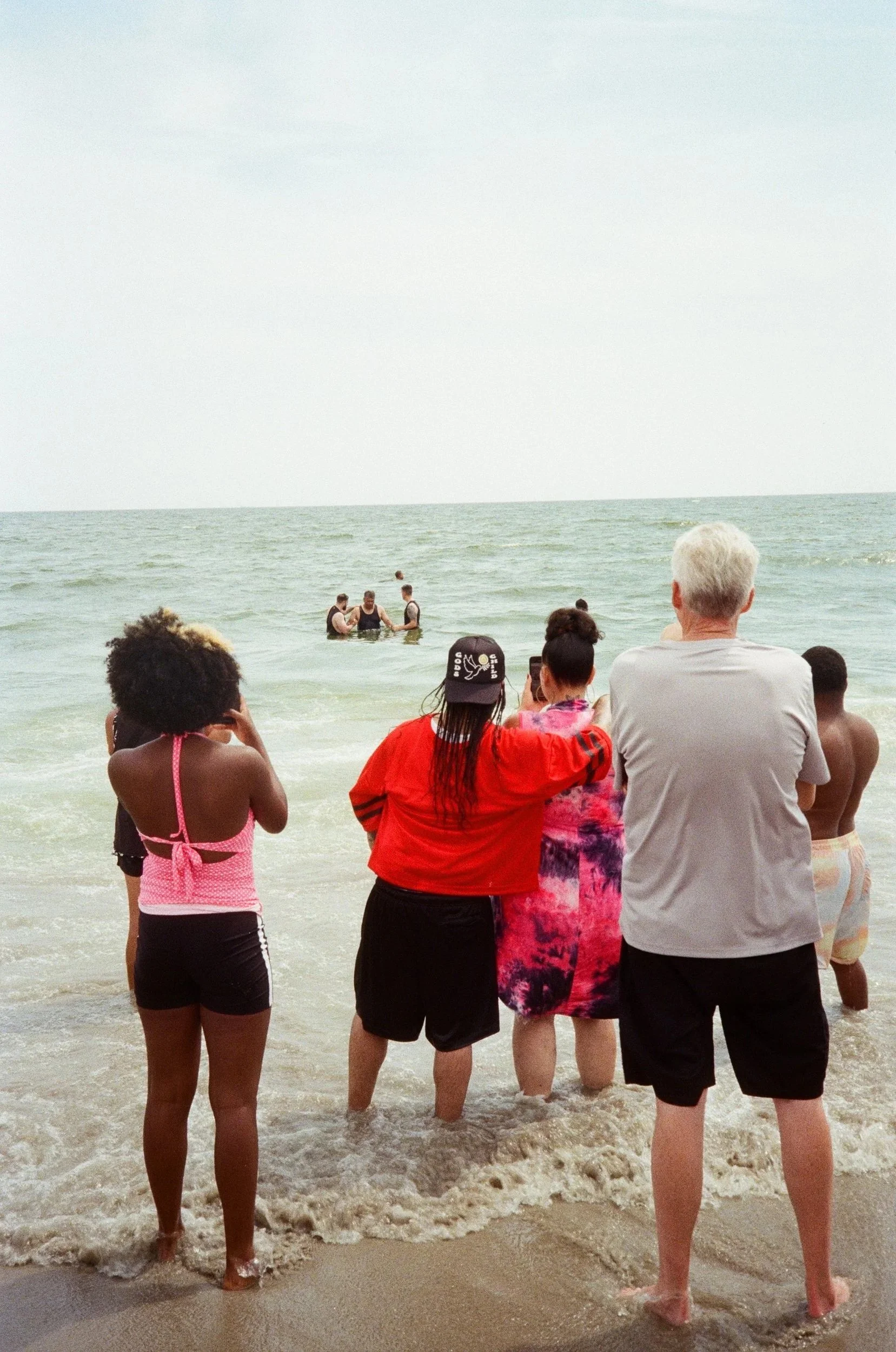 People standing on a beach looking at a small group in the water, with some taking photos.