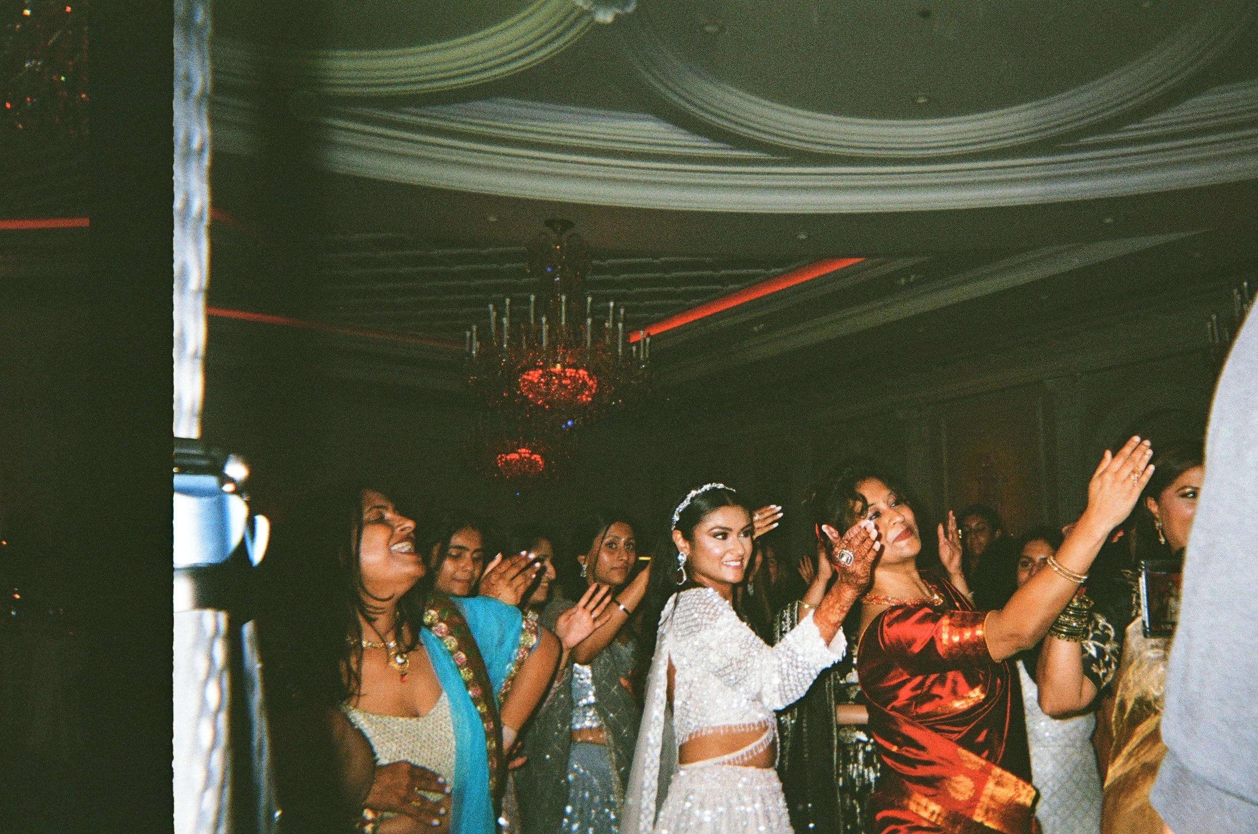 Group of women dressed in elegant traditional Indian attire dancing and celebrating at an indoor event with a chandelier and decorative ceiling.