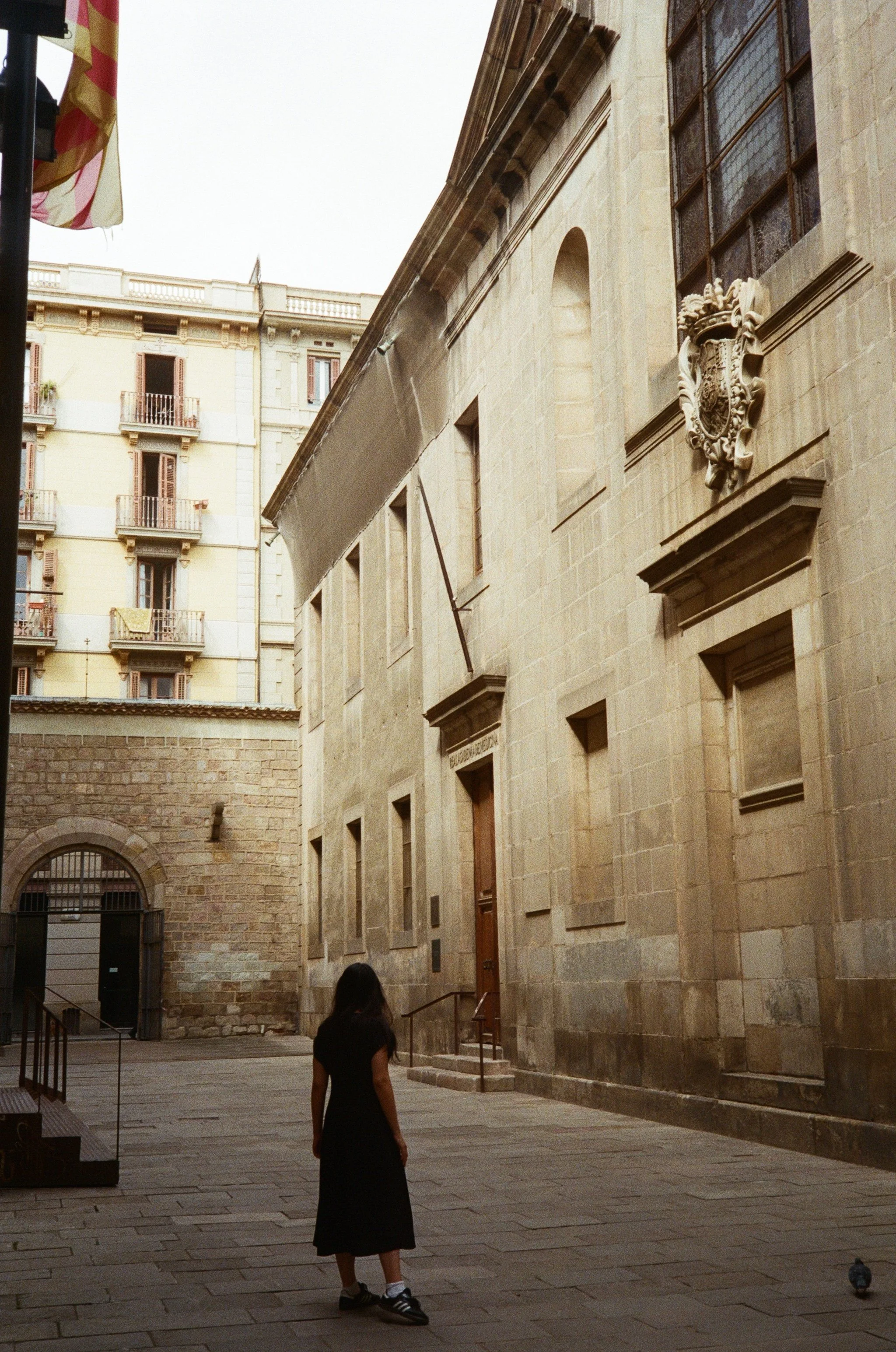 A girl in a black dress and sneakers standing in a narrow courtyard with a historic stone building and taller apartment building in the background.
