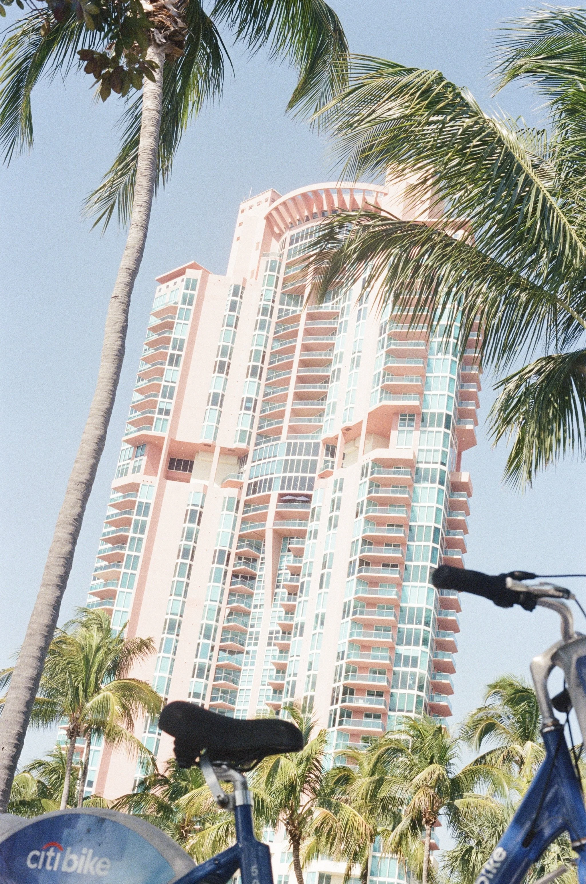 A tall pink and white skyscraper surrounded by palm trees and seen from street level, with a Citibike bicycle in the foreground.