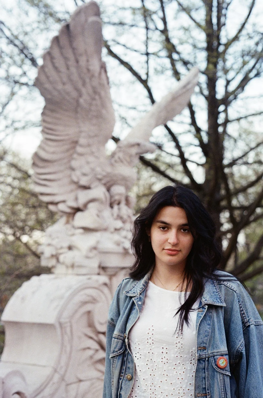 A young woman with long dark hair wearing a denim jacket and a white shirt, standing outdoors with a large white sculpture of an eagle in the background, surrounded by trees.