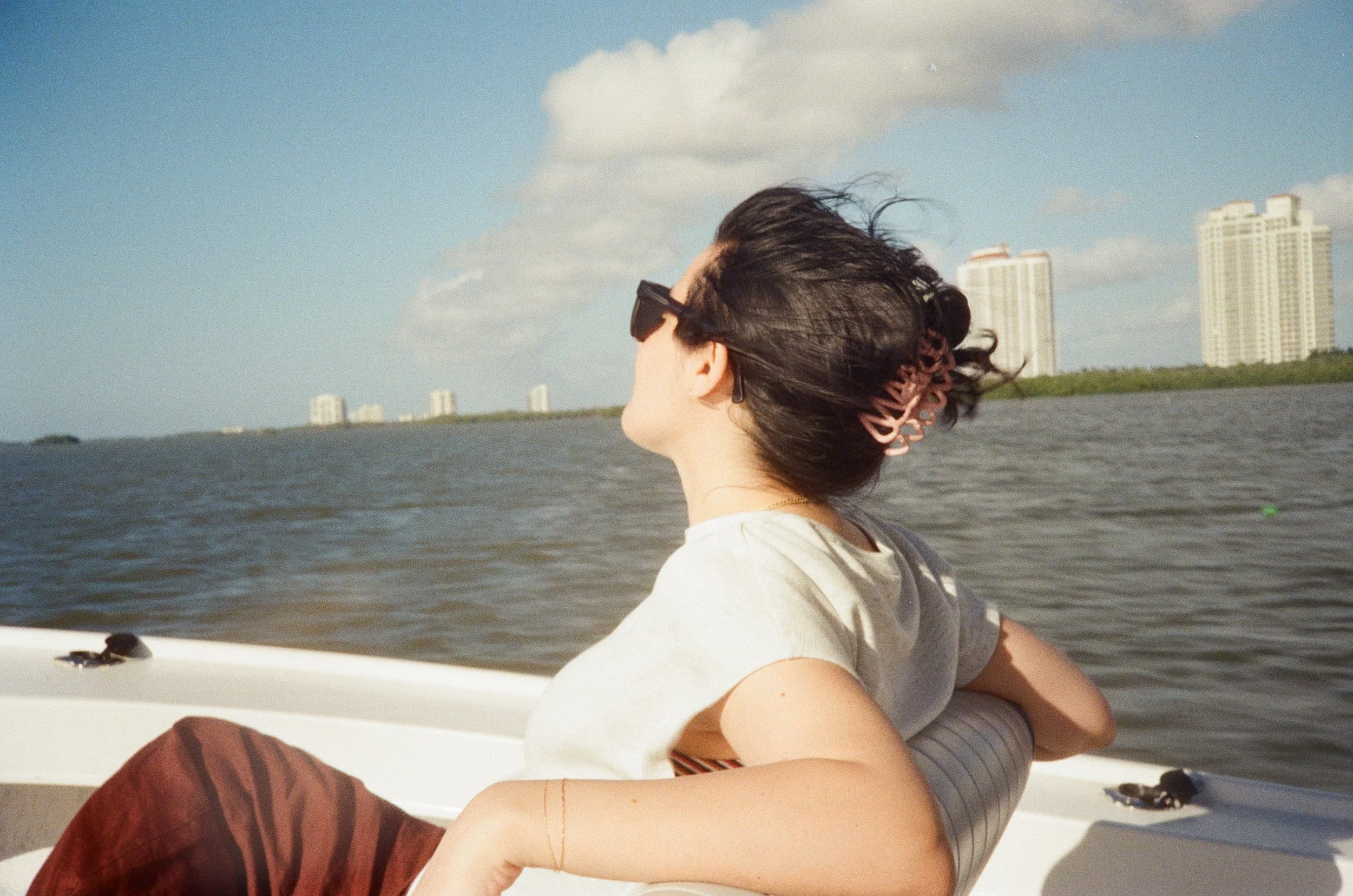 Woman with dark hair in a pink hair clip, wearing sunglasses and a white t-shirt, relaxing on a boat with water and skyline in the background.