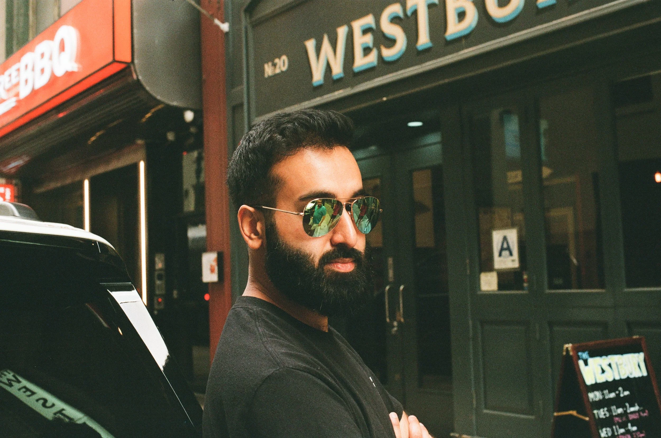 A man with a beard wearing sunglasses and a black t-shirt standing outside a restaurant, with arms crossed, in front of a sign that reads 'WESTBURY'.