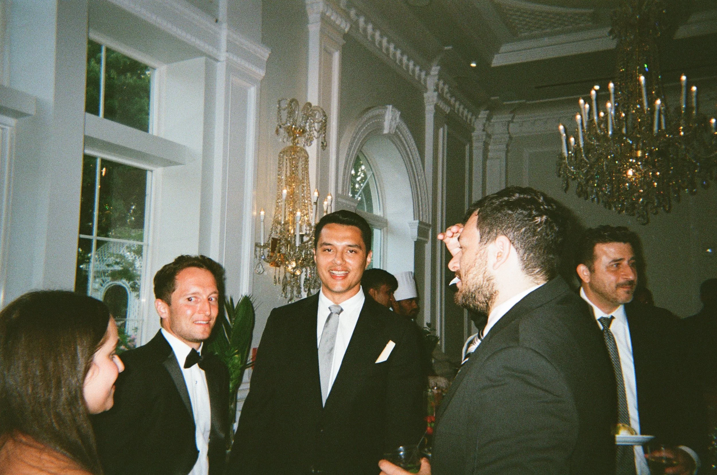 People in formal attire socializing at an elegant event in a decorated room with chandeliers and tall windows.
