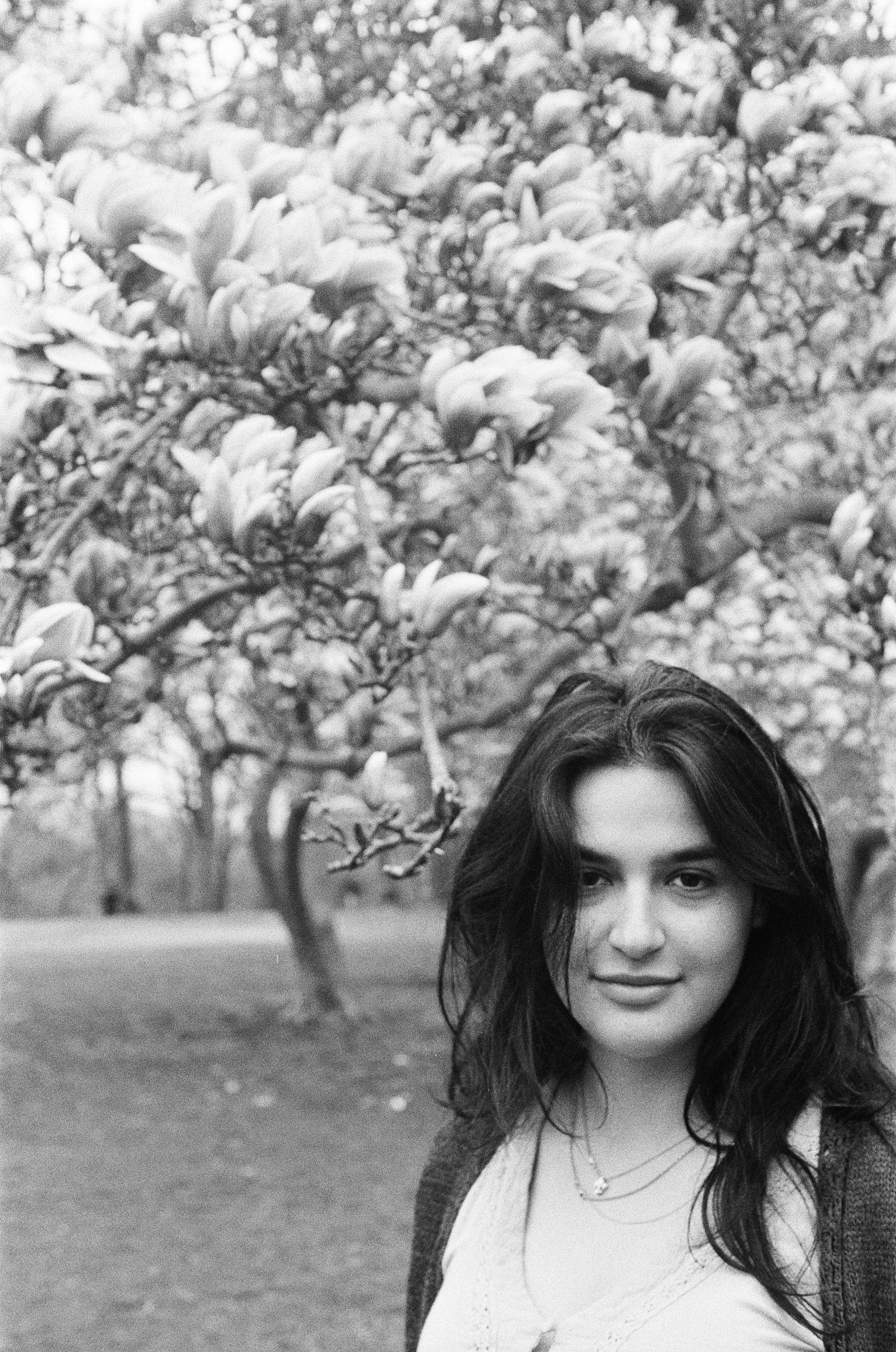 A young woman with dark hair smiling in a park with blooming trees in the background.