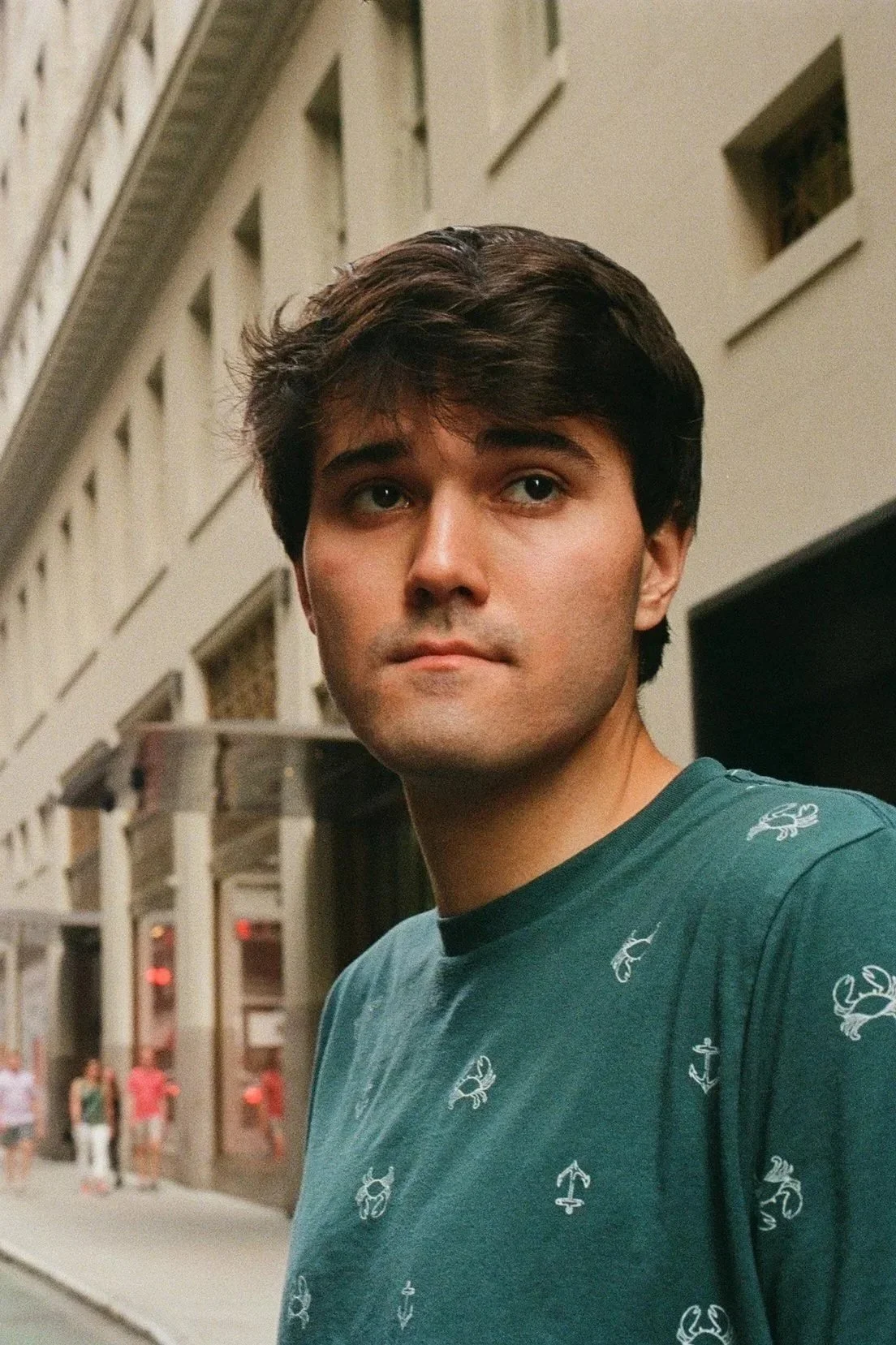 A young man with dark hair and wearing a teal shirt with small white crab and anchor prints, standing on a city street with buildings and pedestrians in the background.