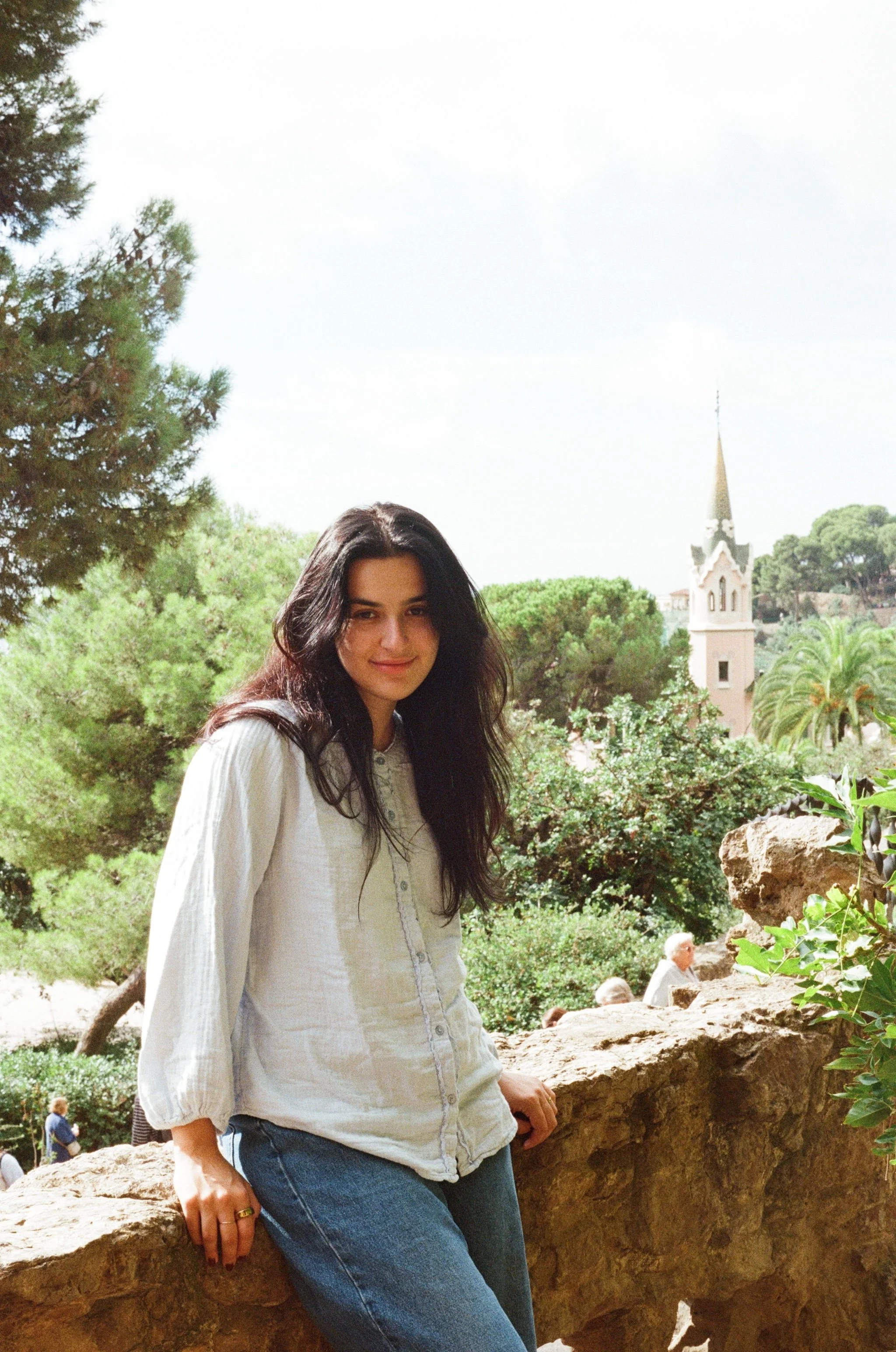 A young woman with long dark hair wearing a white blouse and blue jeans, standing outdoors on a stone wall with a background of trees, a church steeple, and a partly cloudy sky.