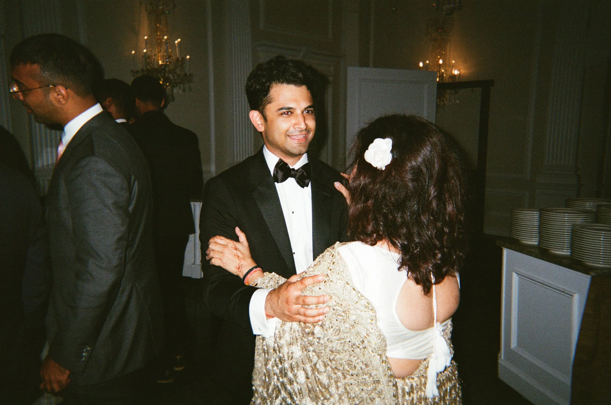 A man in a tuxedo dancing and smiling while looking at a woman with dark hair and a white flower in her hair, in an elegant room with chandeliers.