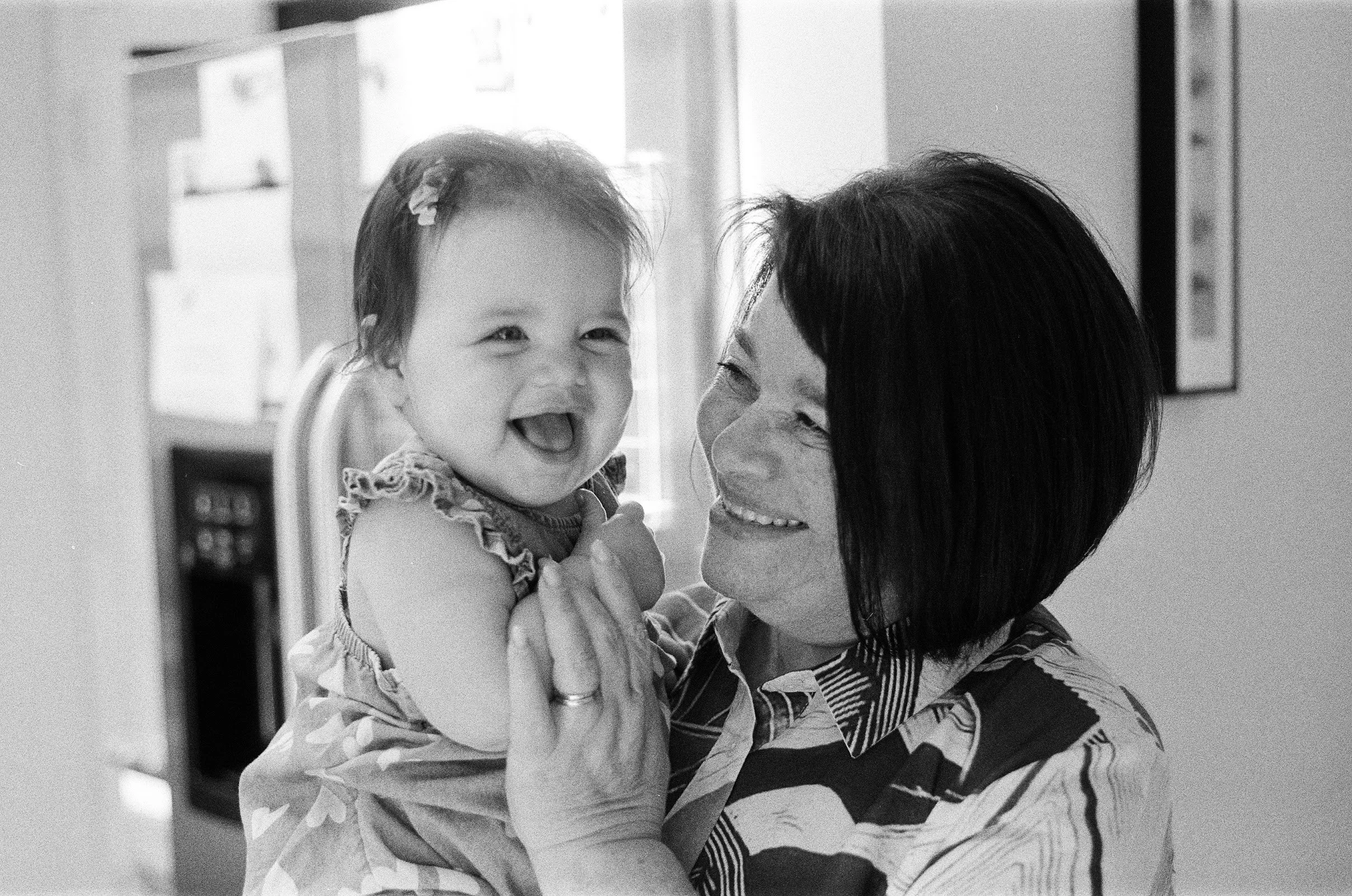 An elderly woman smiling and holding a young girl who is also smiling. They are indoors, with the woman looking at the girl.