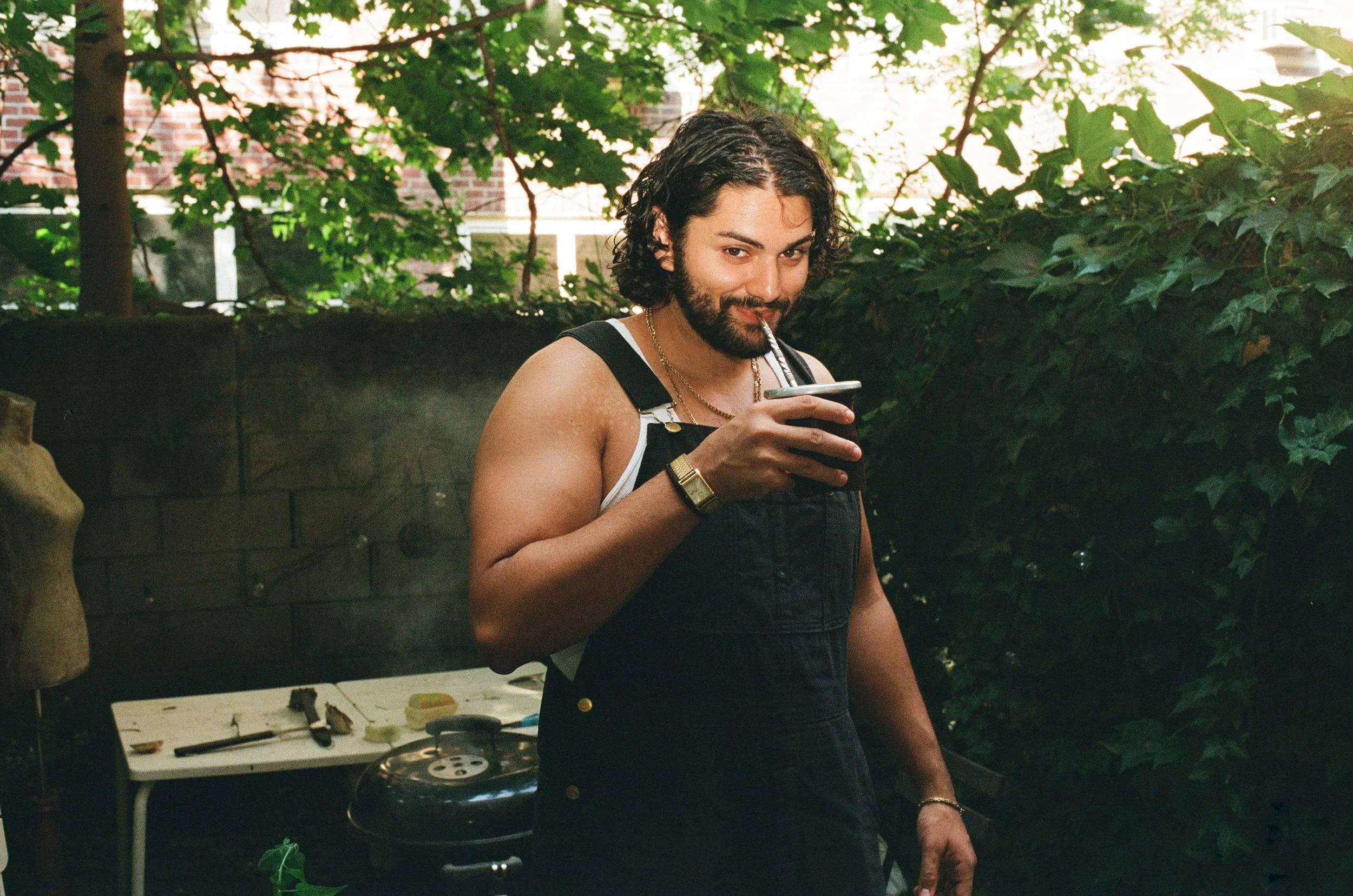 A man with curly dark hair and a beard, wearing black overalls and a watch, drinks a dark beverage from a cup with a straw in an outdoor setting with greenery and trees.