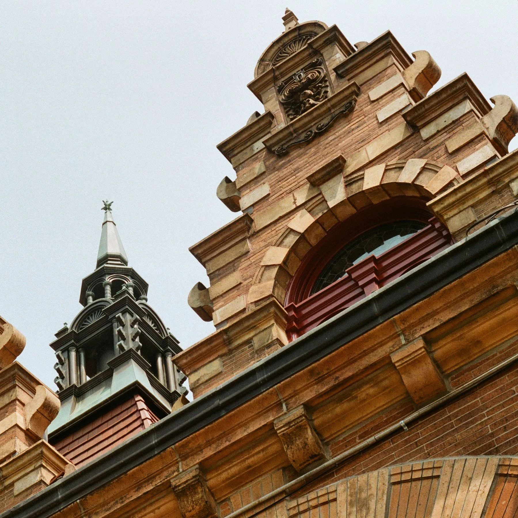 Close-up of historic brick building with ornate architectural details, including clock face, arched window, and decorative elements, against an overcast sky.