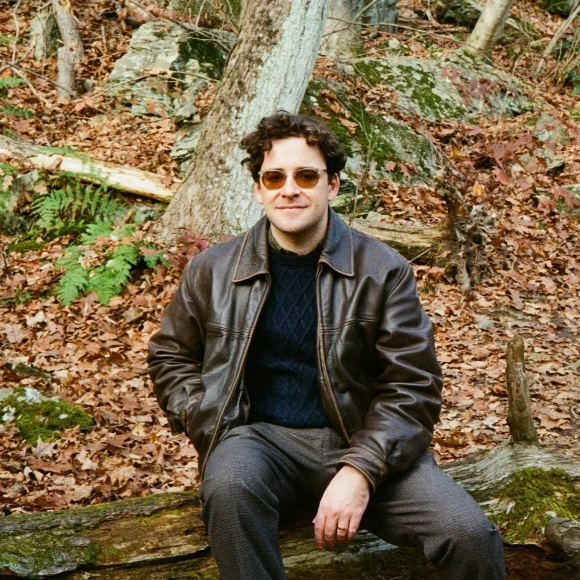 A man sitting on a fallen tree in a forest with autumn leaves, wearing sunglasses, a dark sweater, a leather jacket, and gray pants.