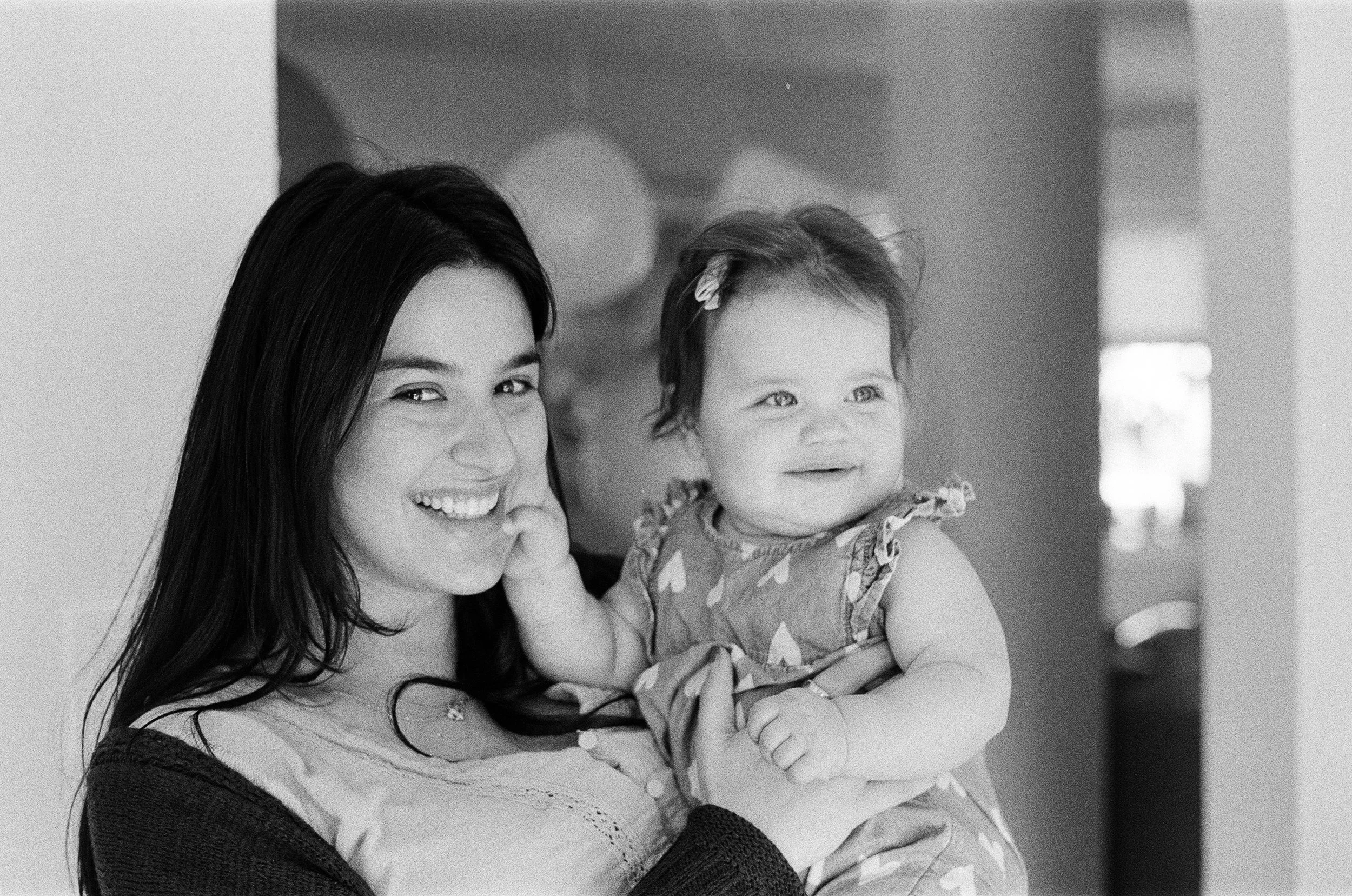 A woman with long dark hair smiling and holding a smiling baby girl with short dark hair, in an indoor setting.