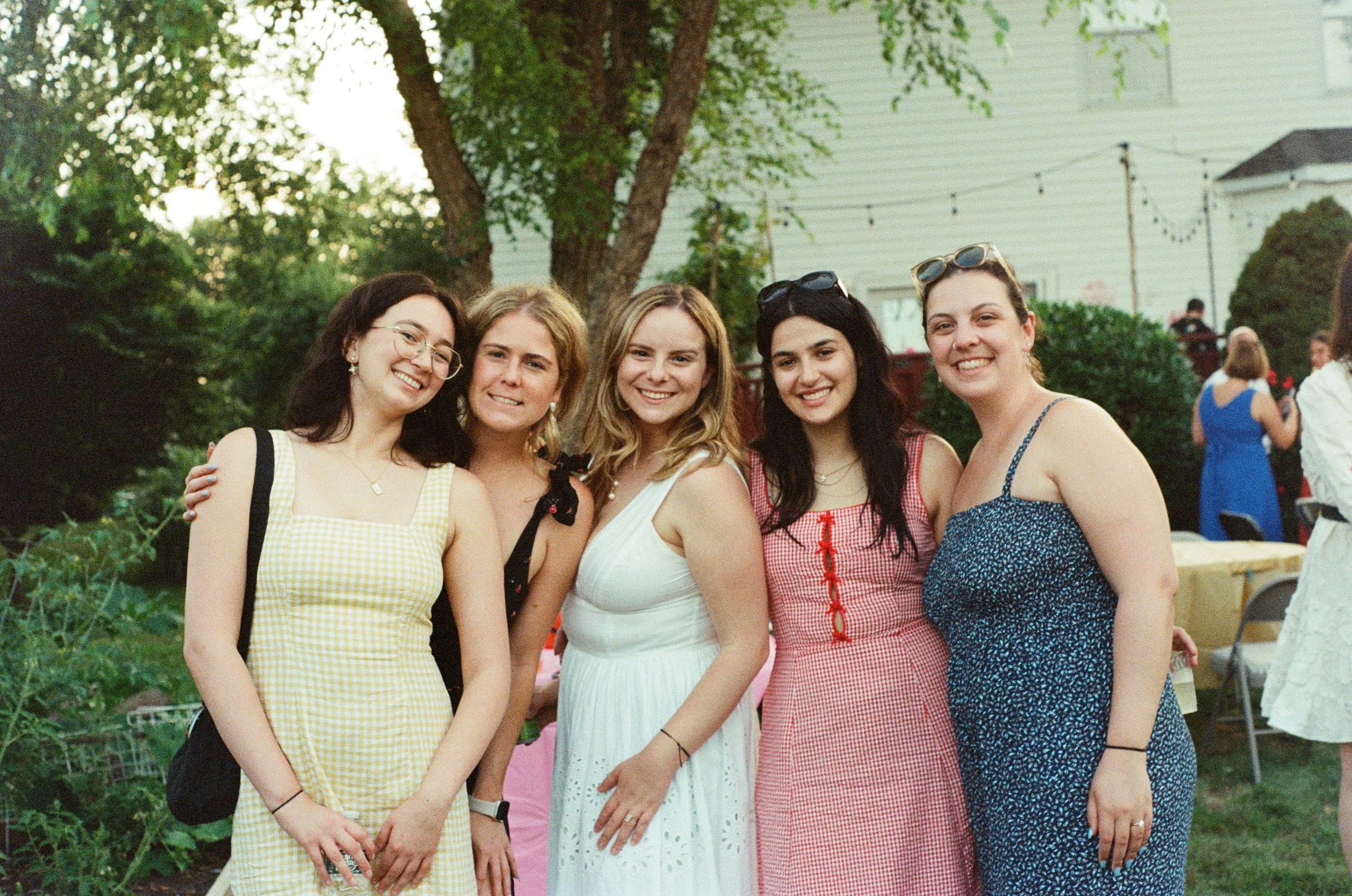 Five women standing outdoors at a gathering, smiling, with trees and a house in the background.