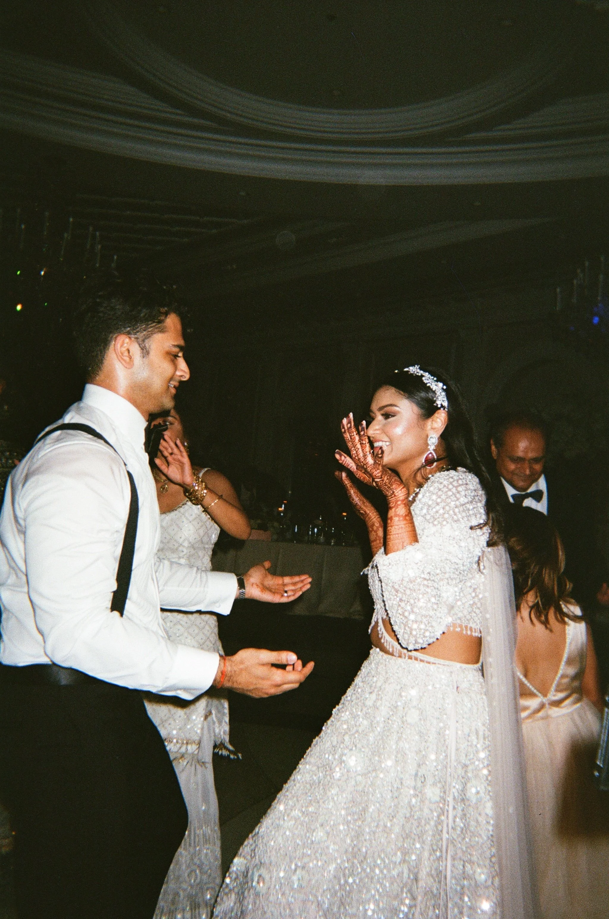 Bride and groom dancing at their wedding reception, with the bride wearing a white embellished gown and the groom in a white shirt with suspenders. The bride is showing henna on her hands and smiling.