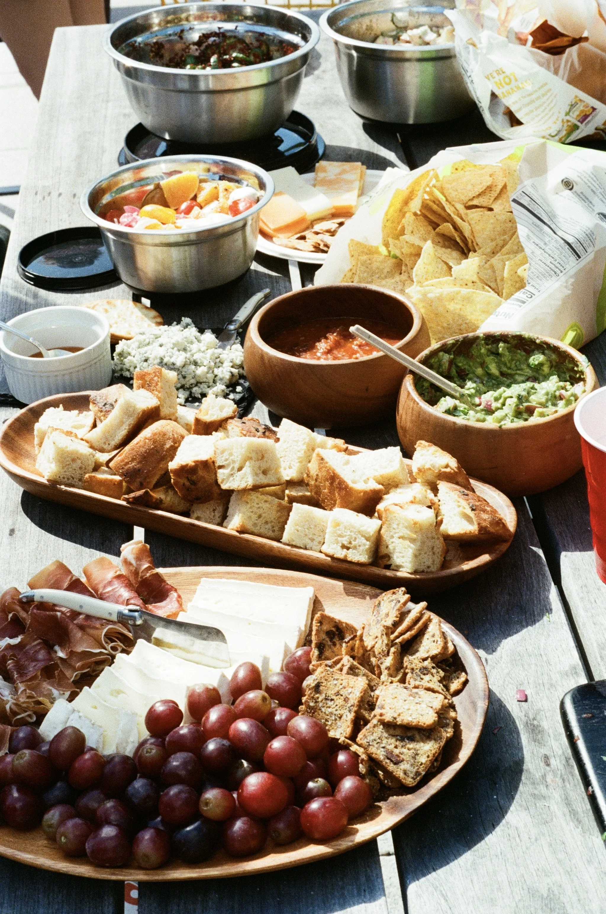 A table with various Mexican food items including bowls of salsa, guacamole, chips, sliced bread, grapes, cheeses, cured meats, and dipping sauces.