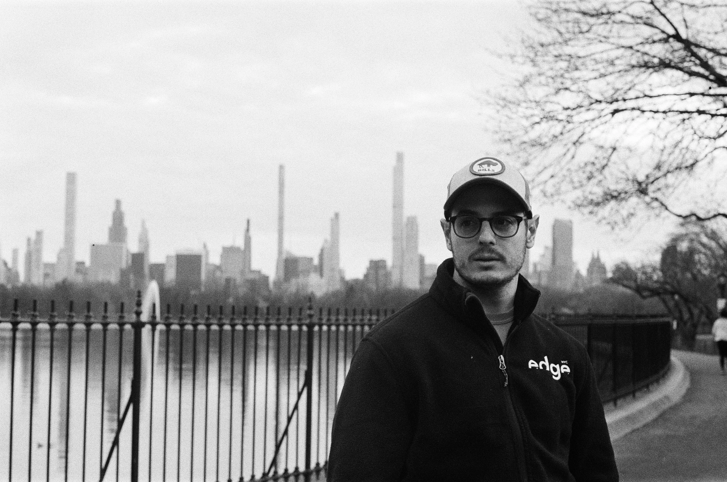 A man wearing glasses, a baseball cap, and a zip-up jacket with the logo 'edge' stands by a black railing overlooking a city skyline in the background, with trees on the right side.