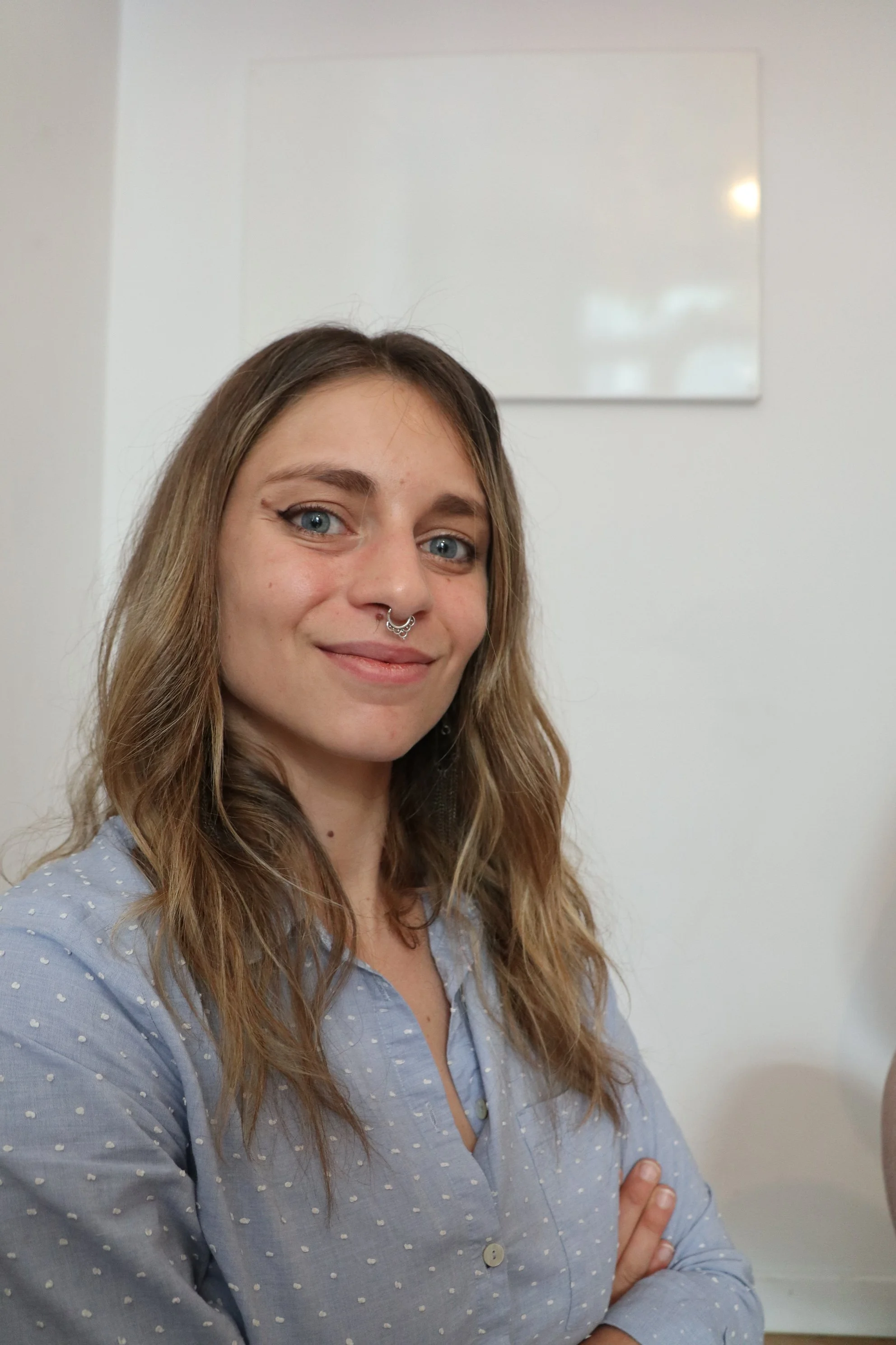Young woman with long wavy hair, wearing a blue button-up shirt with white polka dots, smiling with crossed arms, standing indoors in front of a white wall with a blank whiteboard.