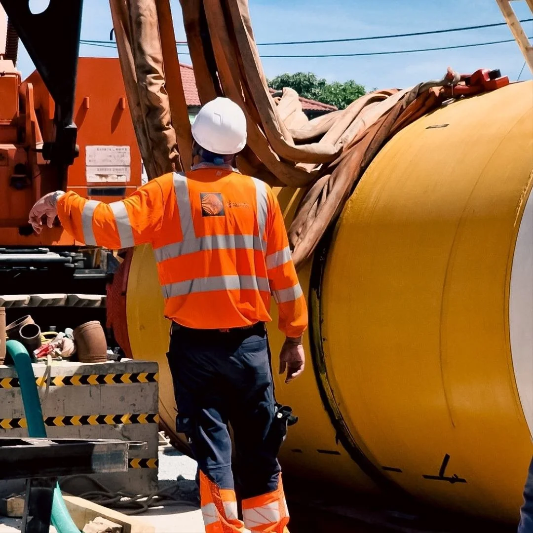 Construction worker in orange safety jacket with "SLS Tunneling" written on the back, wearing a white helmet standing next to a large yellow pipe or cylinder, on a construction site.