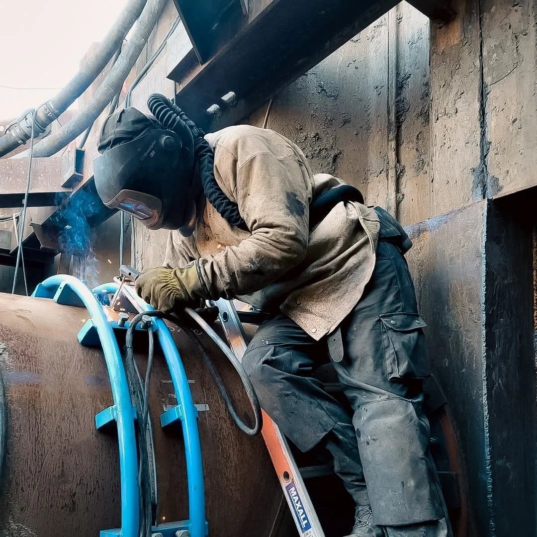 A worker welding on a large metal pipe, wearing protective welding helmet, gloves, and work clothes, with a ladder supporting them.