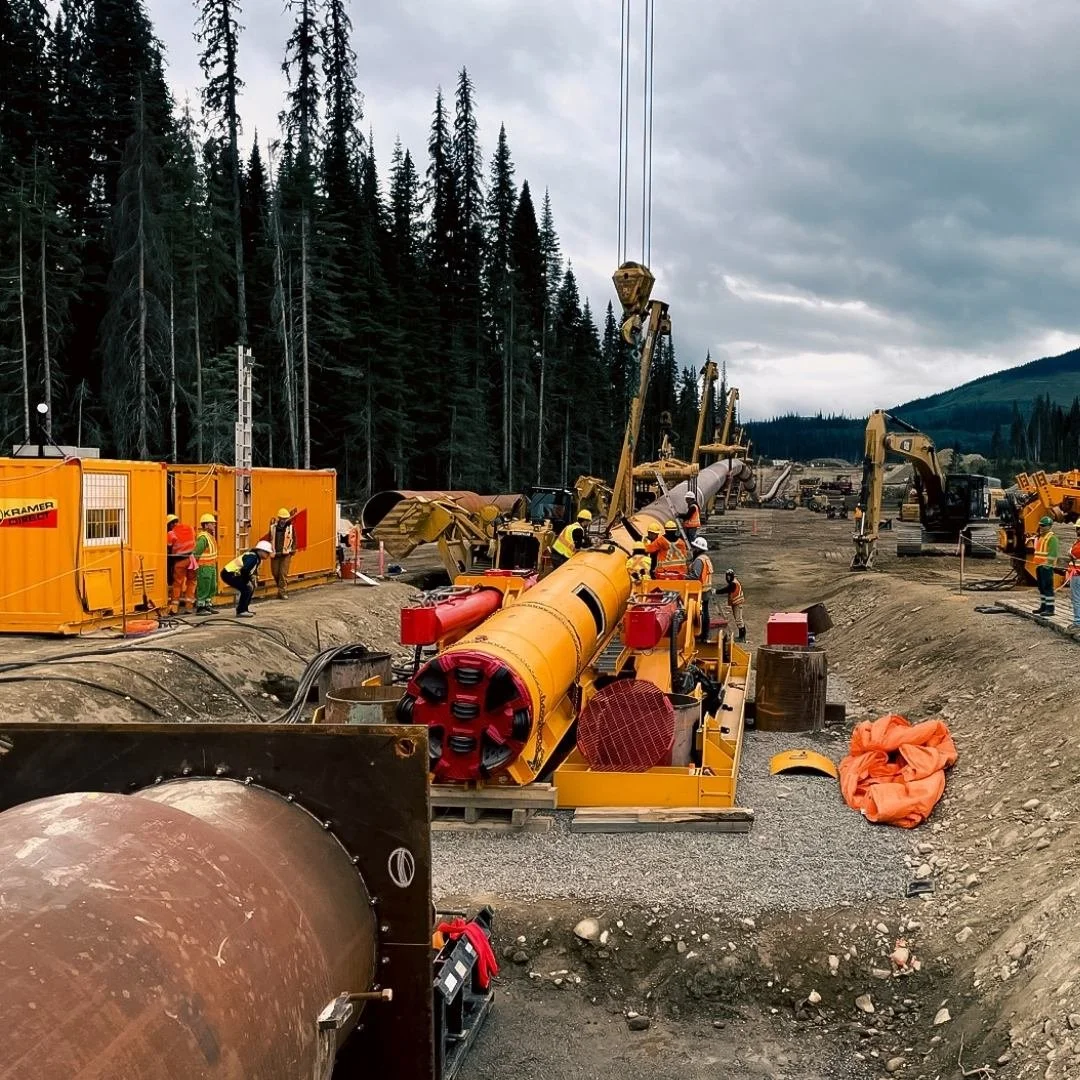 Drone shot of construction workers working on a large infrastructure project outdoors, surrounded by equipment, machinery, and pipelines, with a forested area and hills in the background and cloudy skies overhead.