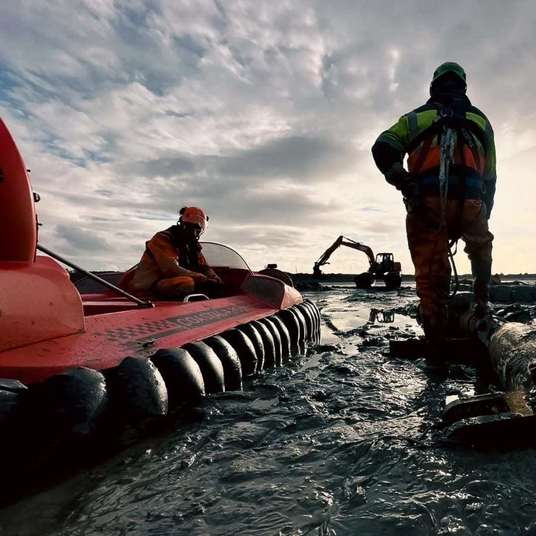 Rescue workers in survival gear working in shallow water near a large vessel with a logo, using equipment like pipes and machinery under a cloudy sky.