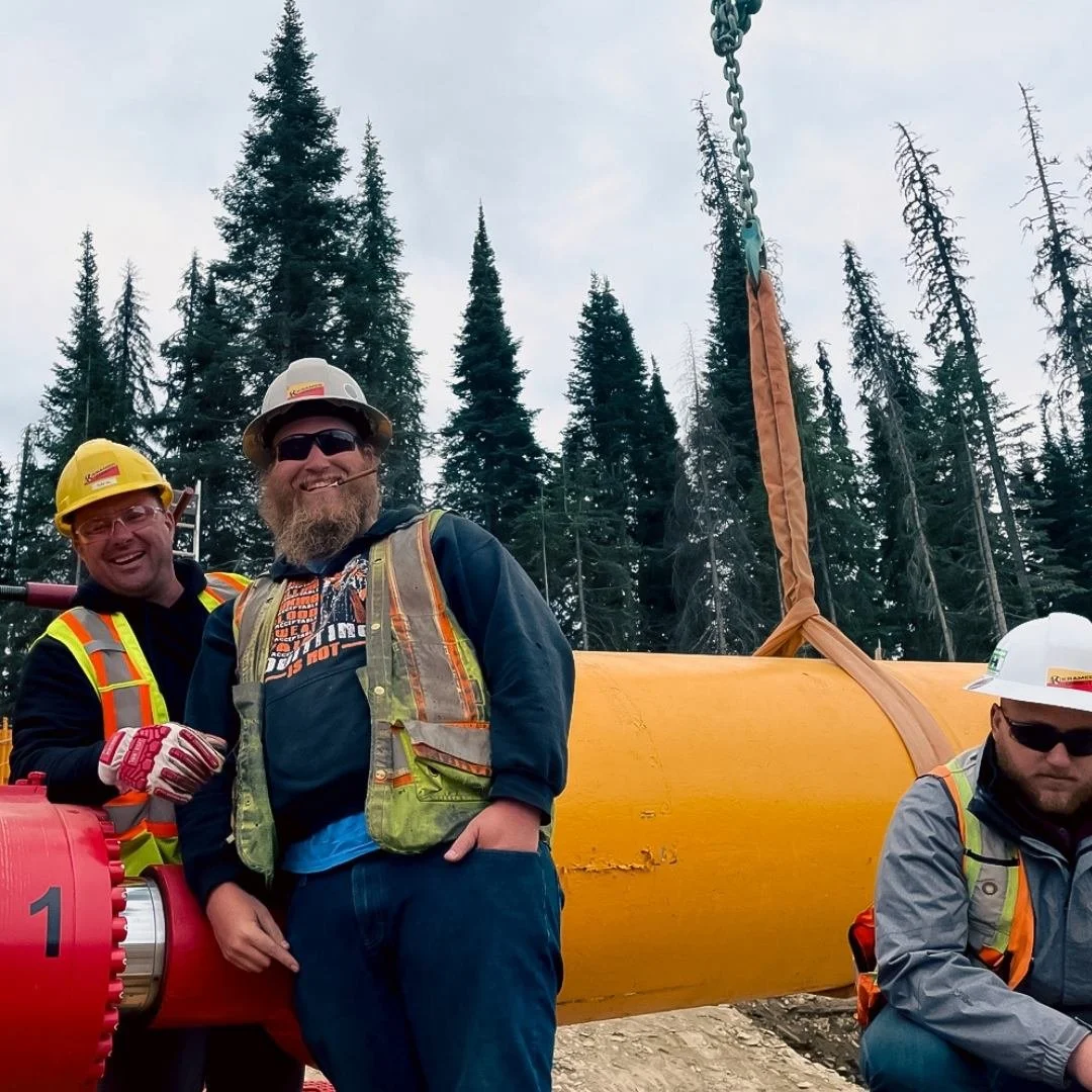 Three construction workers in safety gear stand in front of a large yellow pipeline in a forested area, smiling at the camera - holding a sign that reads "excellent brand and venture partner"