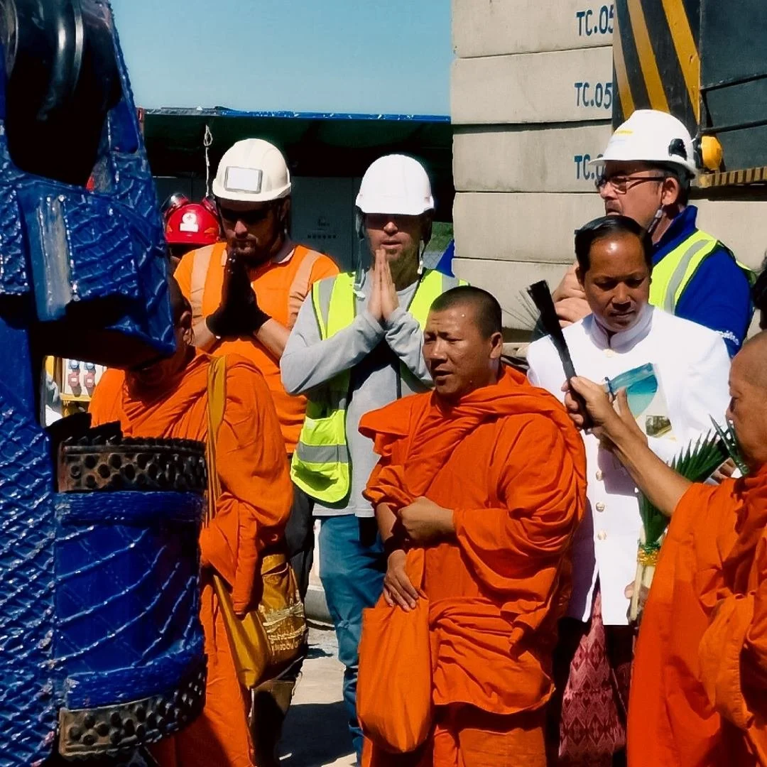 Group of Buddhist monks in orange robes and construction workers in safety gear praying outdoors, with a professional videographer from Doncaster partially visible in the background.