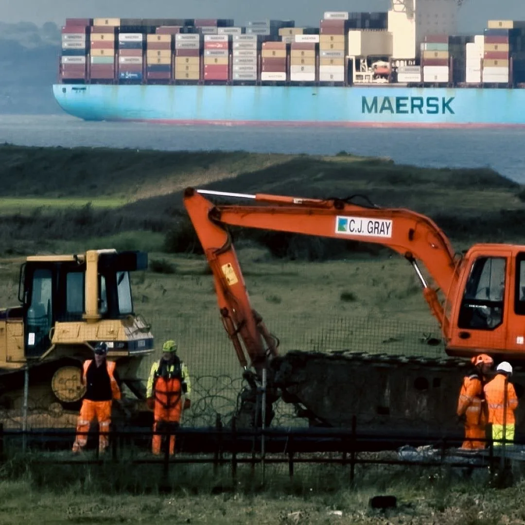 Construction workers in orange safety gear and helmets operating heavy machinery on a construction site near a fenced area, with a large commercial container ship labeled 'MAERSK' in the background on the water.