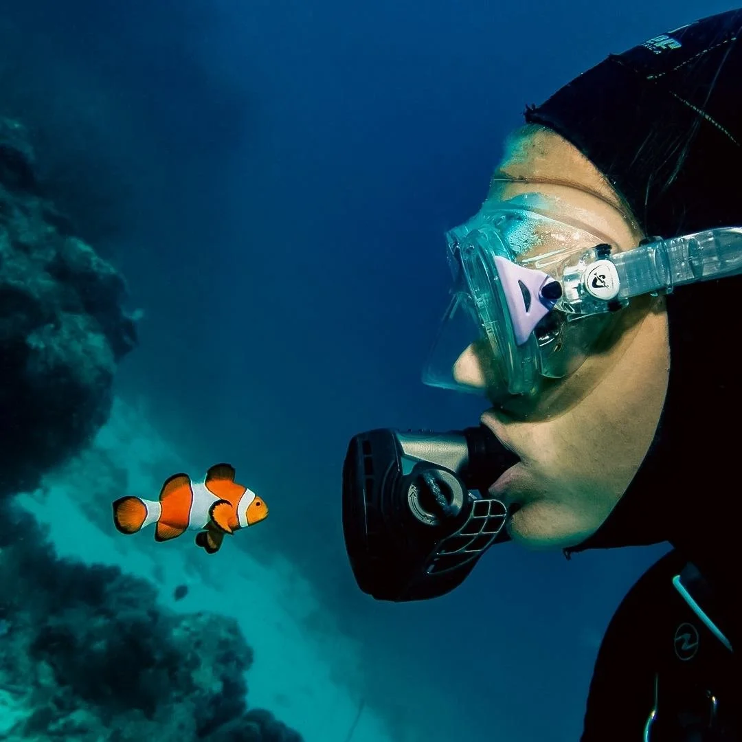 Person wearing a diving mask and snorkel exploring underwater near a clownfish and coral reef.