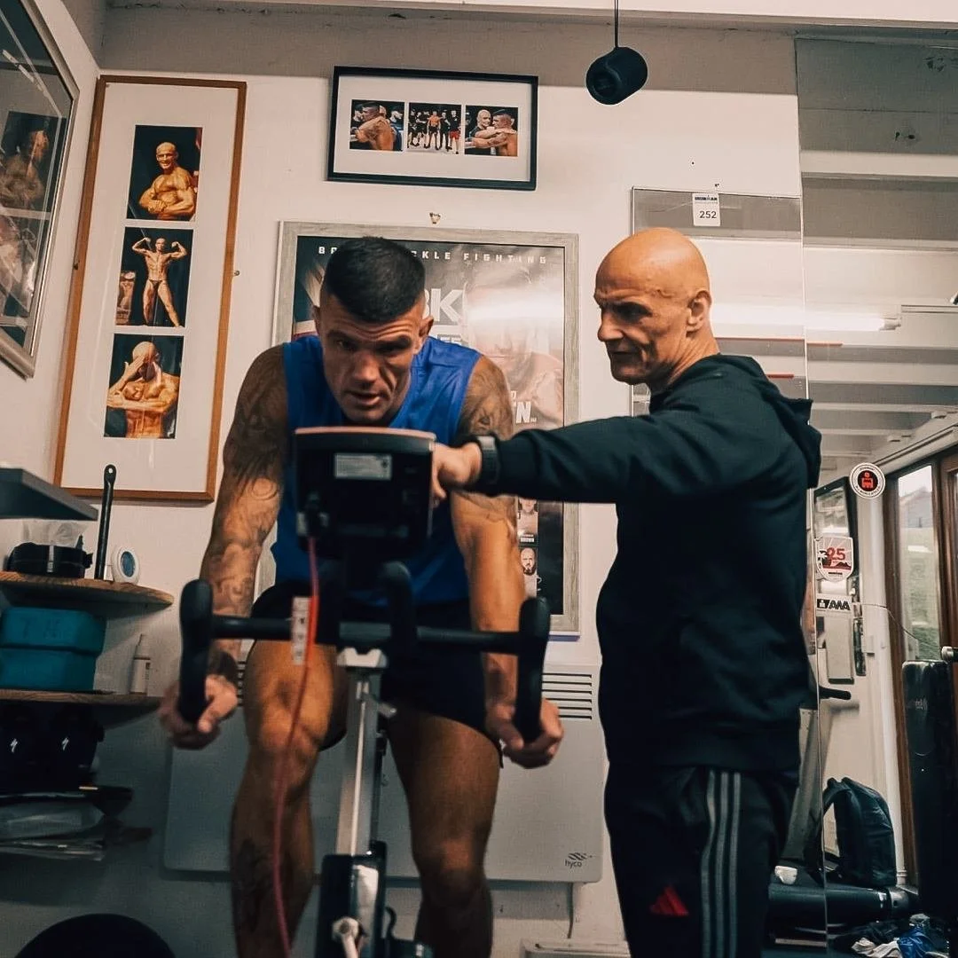 Professional fighter in blue tank top is riding a stationary exercise bike, supervised by a trainer in black attire inside a gym. The background includes framed posters and photographs on the wall.