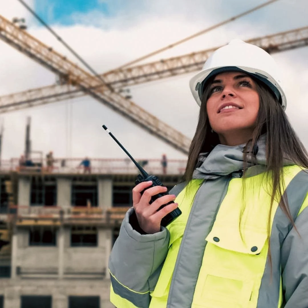 A female construction worker from Doncaster with long brown hair, wearing a white safety helmet and a high-visibility yellow safety vest, holding a walkie-talkie at a construction site with cranes and a building under construction in the background.