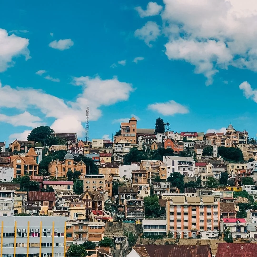 A drone shot of a cityscape Madagascar with numerous houses and buildings on a hillside, with a blue sky and scattered clouds overhead - with text overlaid "drone services in the UK"