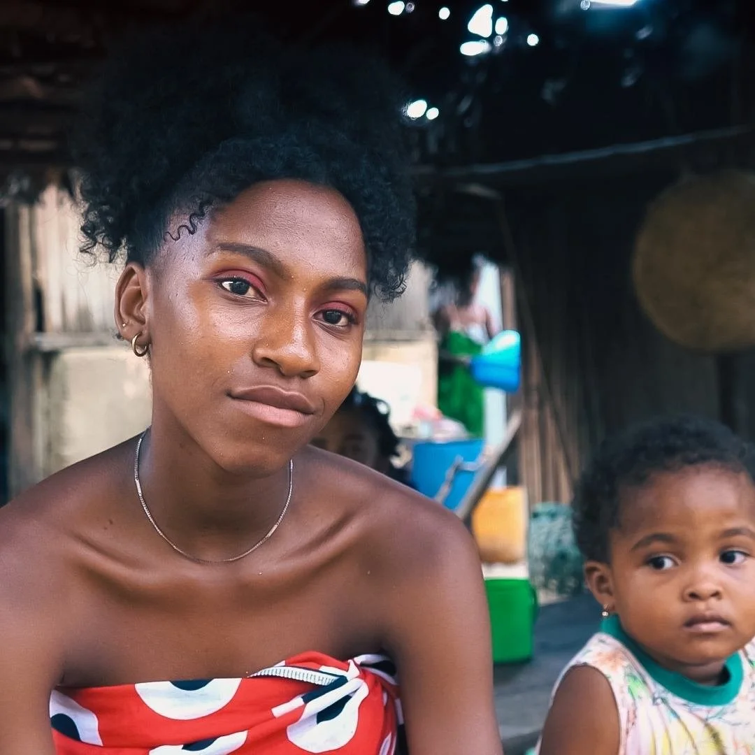 A woman and a young girl sitting together inside a wooden hut in Madagascar, both with short curly hair and wearing a strapless dress - and another young girl - with a videographer from Soul Revolution festival partially visible in the background