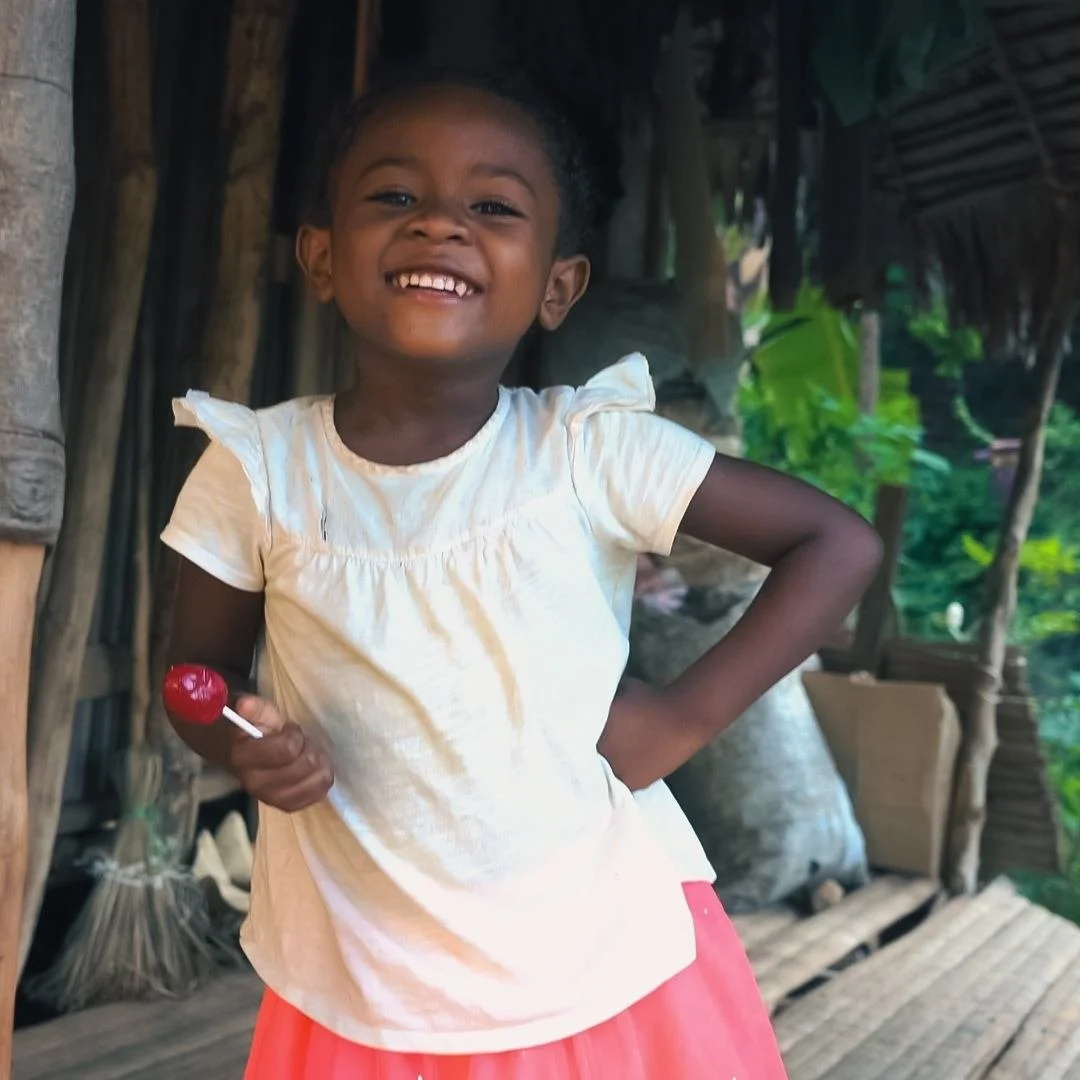 A young girl with dark hair, wearing a white top and pink skirt, standing inside a wooden shack with a thatched roof, smiling and holding a red lollipop - talking to a videographer from Soul Revolution festival