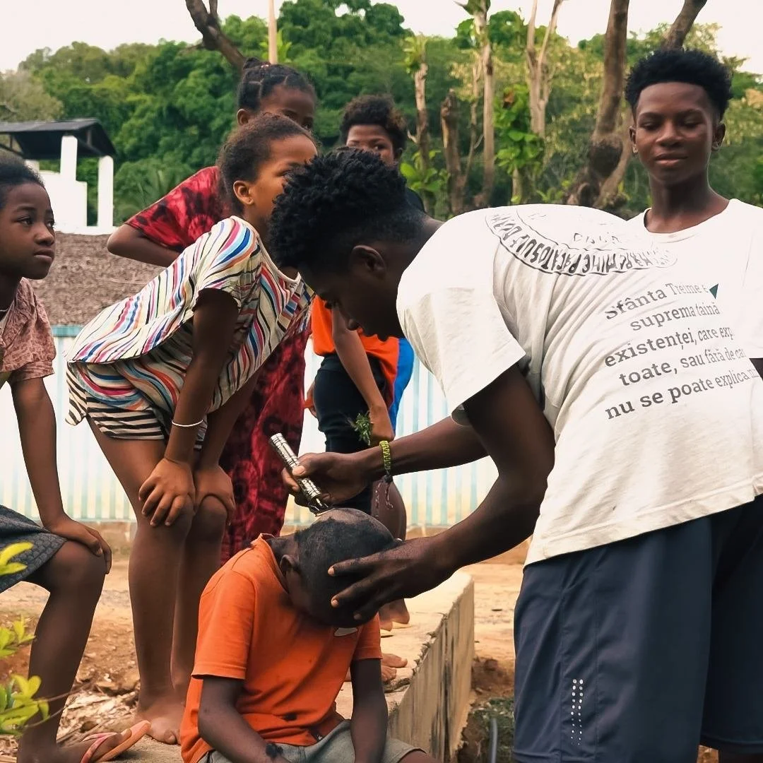 A group of children in Madagascar, gathered around a man who is giving a haircut to a seated boy during the daytime outdoors - with a videographer from Soul Revolution festival visible in the background.