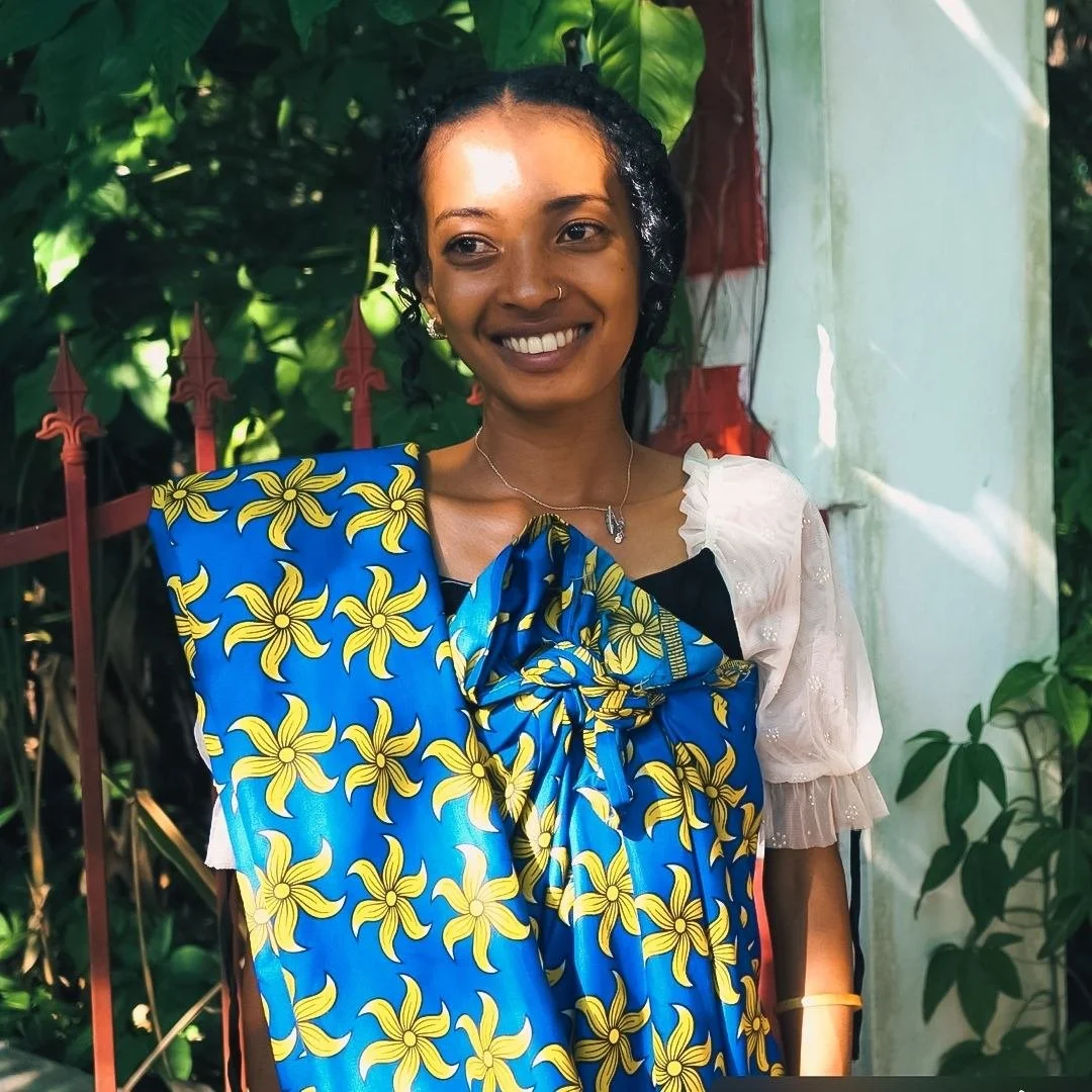 A Madagascan woman with dark curly hair, gold earrings, and a necklace, smiling and standing outdoors near green foliage. She is wearing a colorful traditional dress with yellow floral patterns on a blue background and a white sleeve detail.
