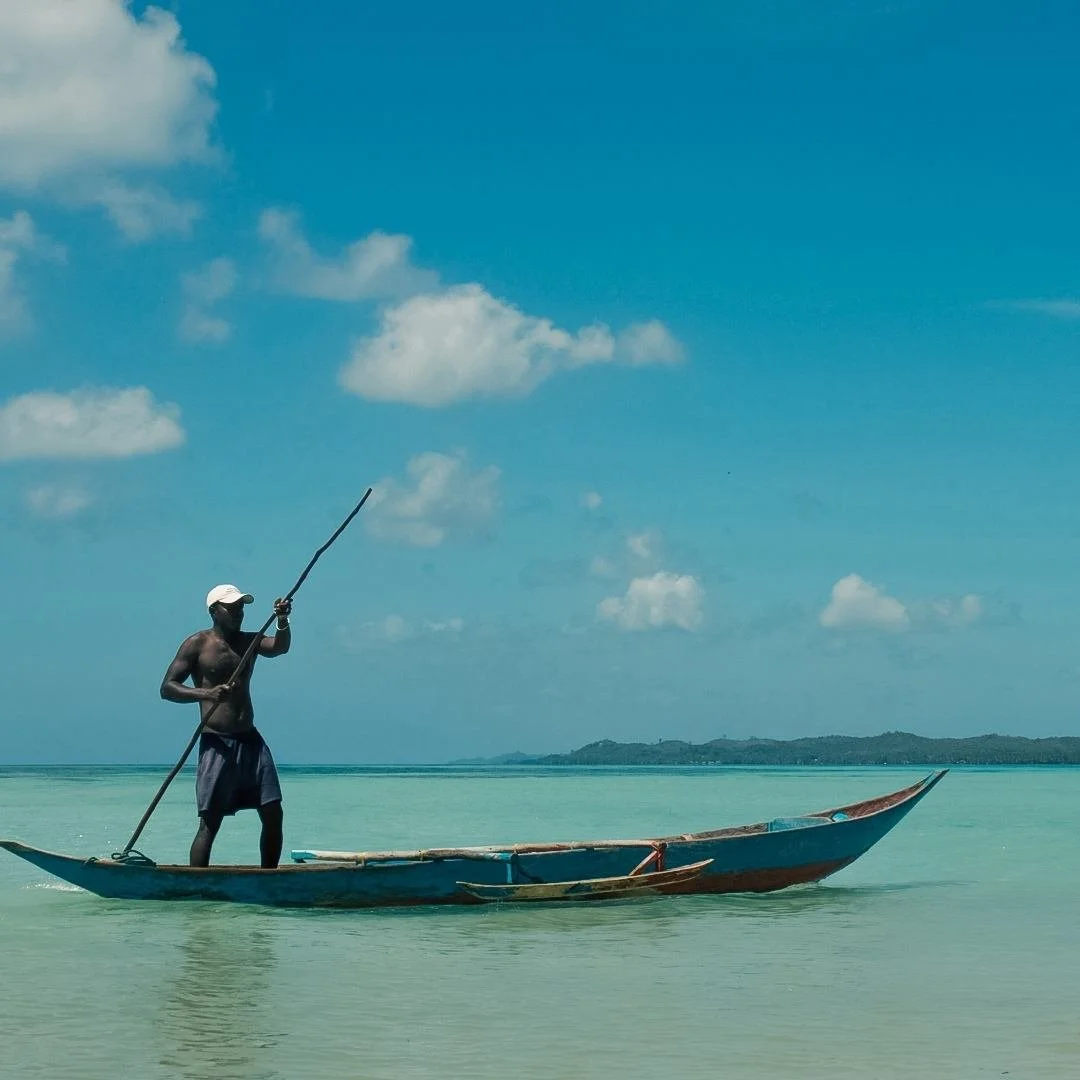 A man paddleboarding on calm, light blue water with a large boat, wearing a white cap and shorts, under a partly cloudy sky.