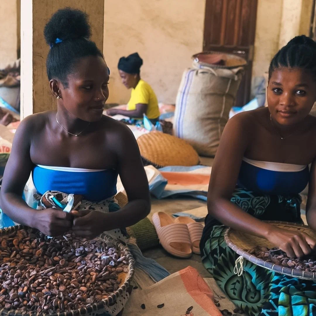 Two women are sitting with baskets of nuts. One woman is wearing a blue dress and the other has a patterned fabric wrapped around her waist. In the background, two videographers from Soul Revolution & Made by UNO