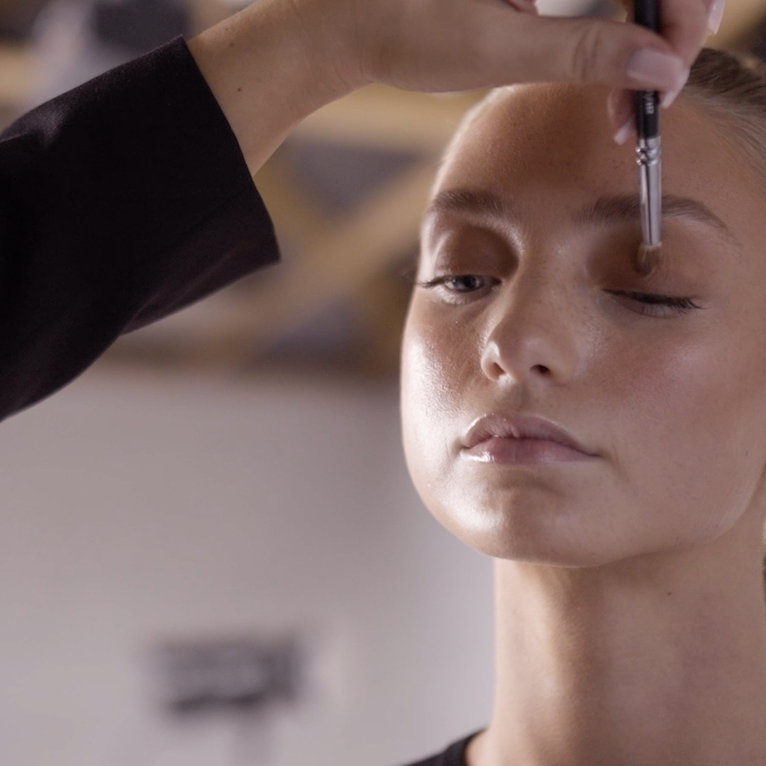 A model during BTS in a Creative Quarter photoshoot - with her eyes closed as a makeup artist applies eyeshadow with a brush.