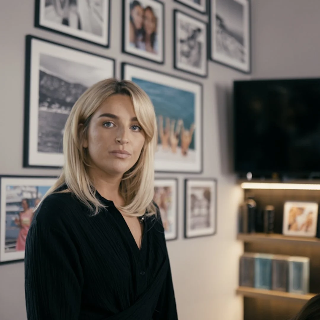 A woman with blonde hair wearing a black top, standing in a room with a gallery wall of framed photographs behind her.