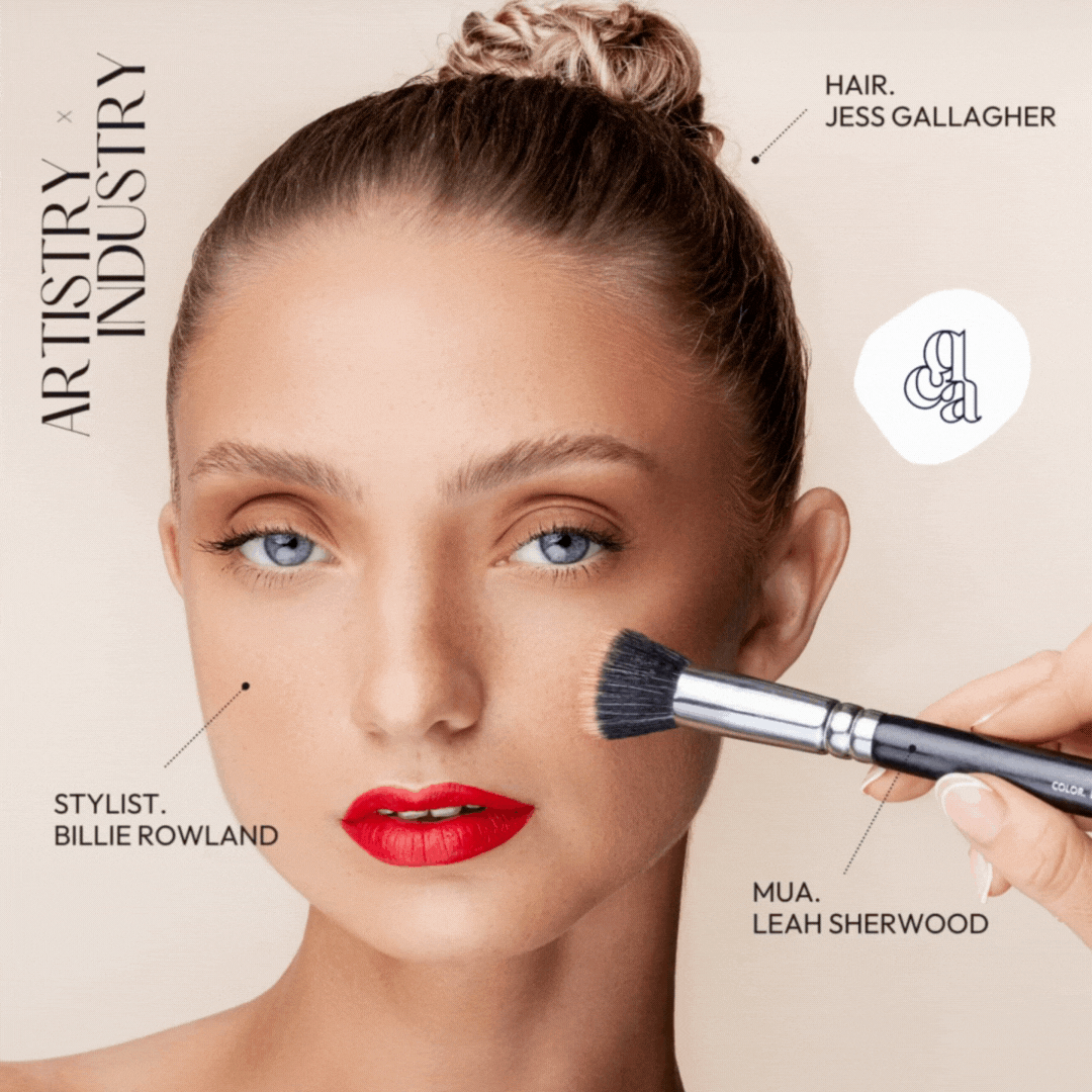 A Model with blue eyes and red lipstick has makeup applied with a brushby her stylist, Billie Rowland of creative quarter, and her makeup artist Leah Sherwood. The text on the image says 'Artistry x Industry' & 'brand by UNO'