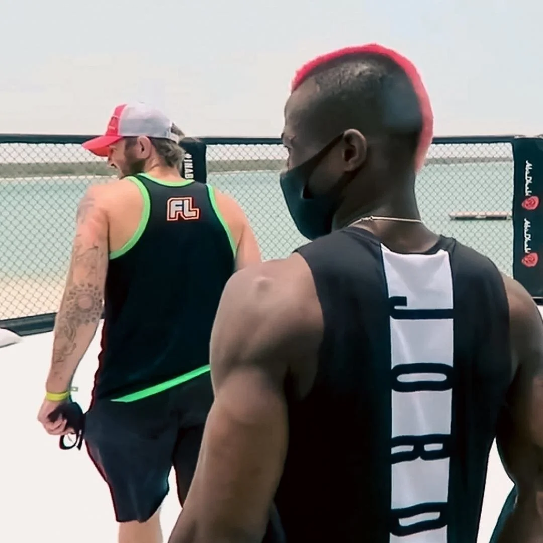 Marc Diakiese and another man in the outdoor ufc octagon on yas island,  wearing a face masks and a sleeveless shirt, the other man with tattoos and wearing a cap with a docuemntary film maker in the background.
