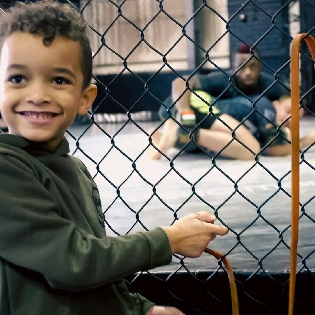 A young boy smiling, looking into an MMA octagon, where his father Marc Diakiese is training inside with Jack Grant