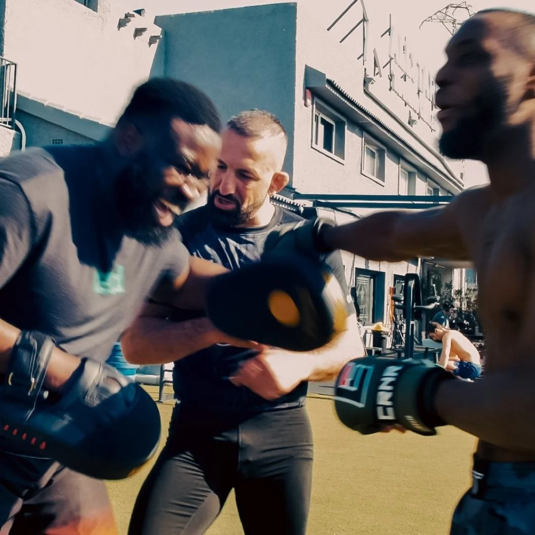 Three men practicing boxing outdoors, one wearing boxing gloves, and the other two holding focus mitts, in the courtyard of All Stars Training centre in Alicante, Spain