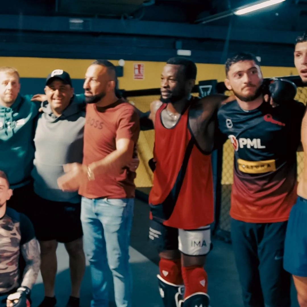 A group of six men standing together with their arms around each other inside a MMA facility. Some are dressed in athletic gear, and one is wearing boxing or MMA gear.