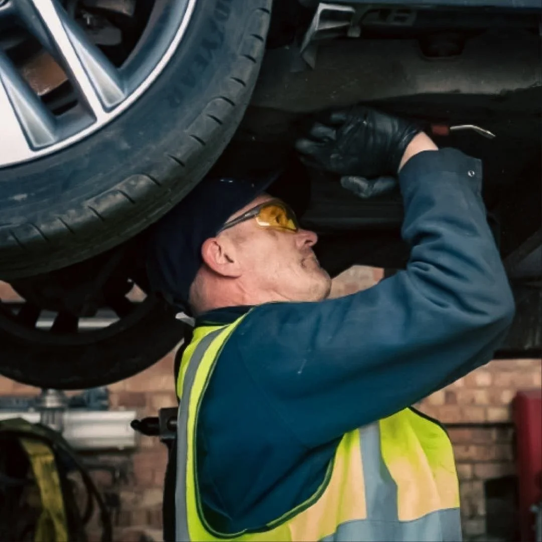 A Synetiq mechanic working underneath a car, wearing safety glasses and gloves, in a garage with a brick wall background.