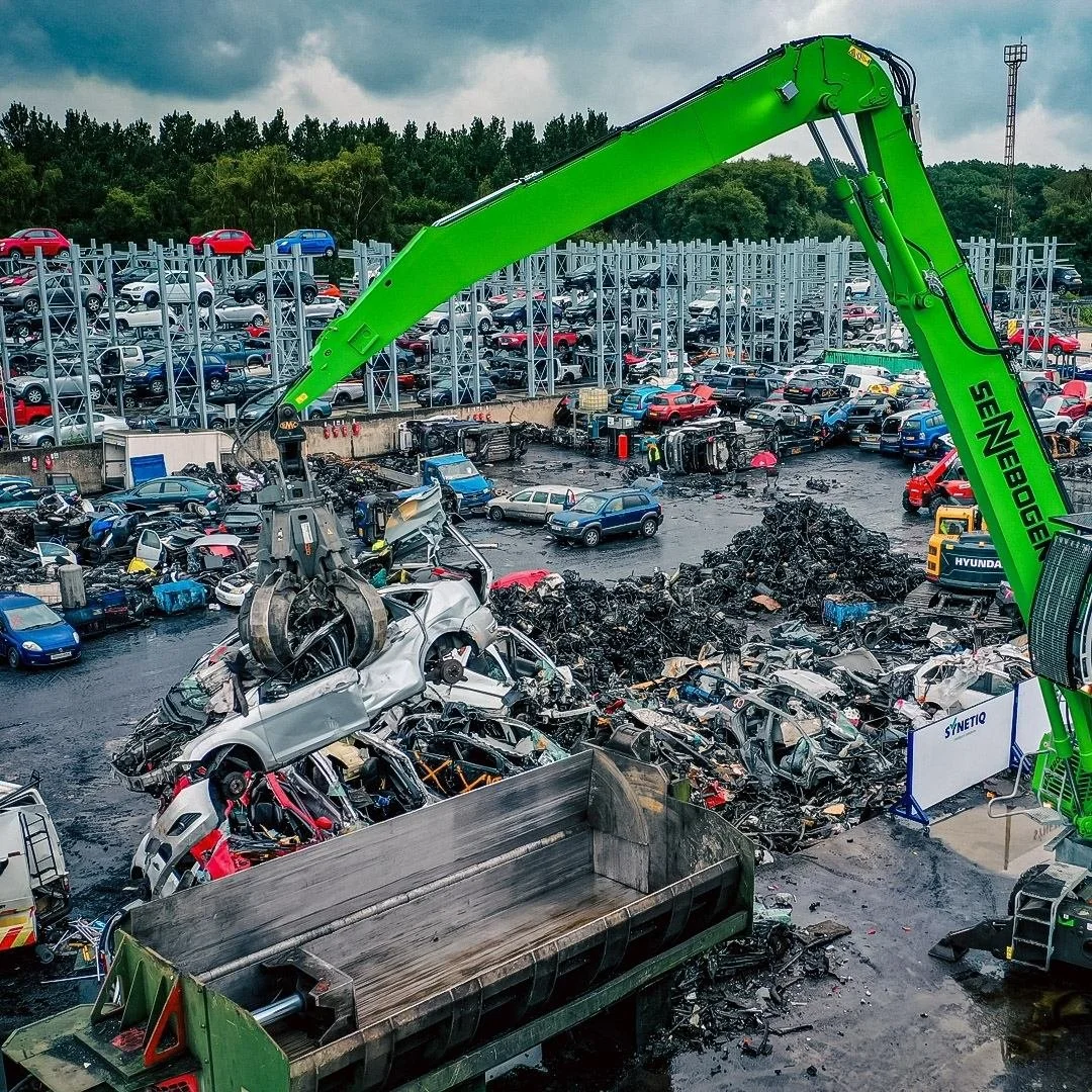 Drone shot of a large green crane lifting wrecked cars at a Synetiq car parts facility in doncaster, with numerous damaged vehicles and scrap metal piled around under cloudy skies.